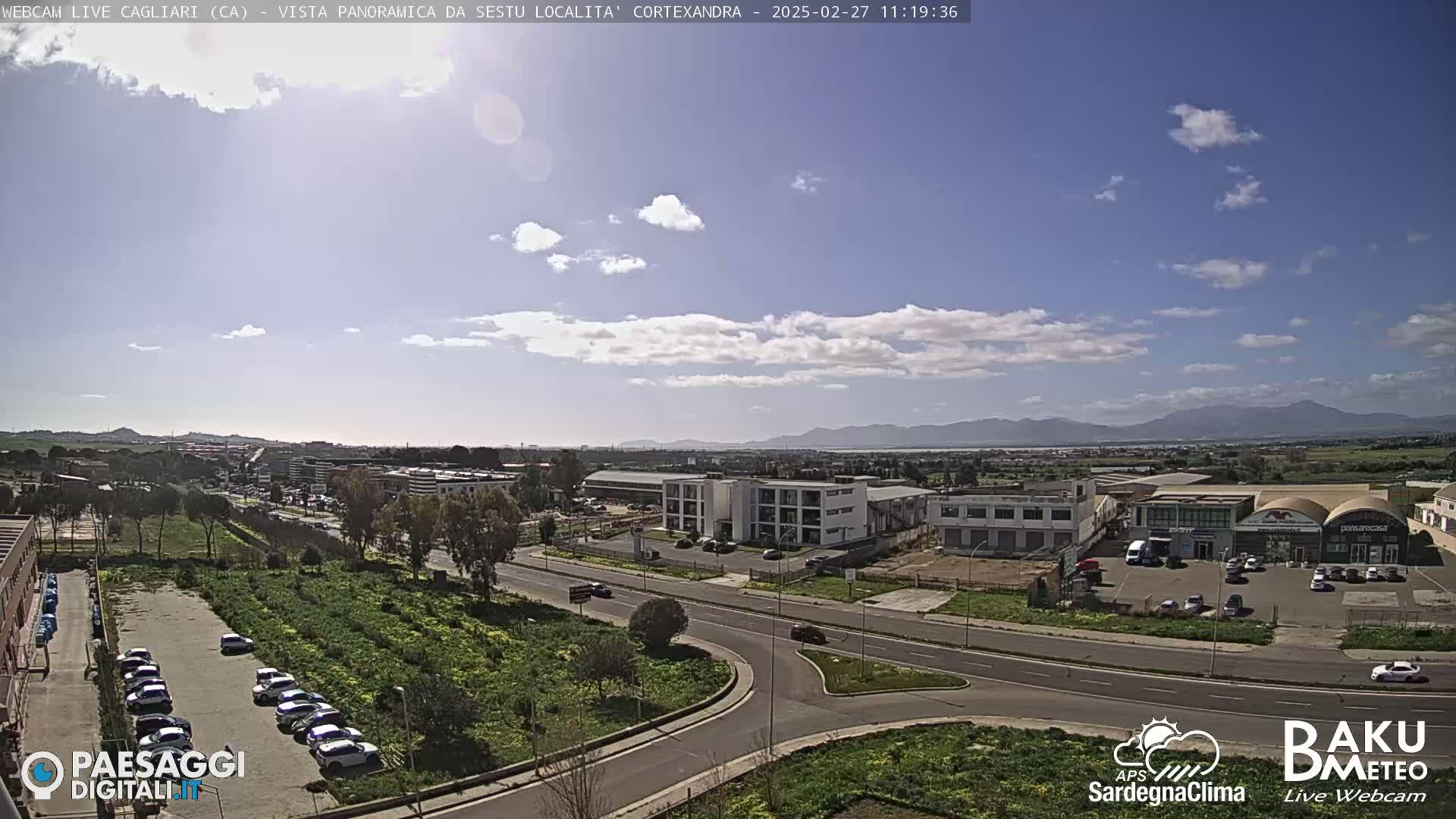 A panoramic view of a city landscape under a partly cloudy sky shows a road intersection, buildings, and parked cars, with mountains visible in the distance.