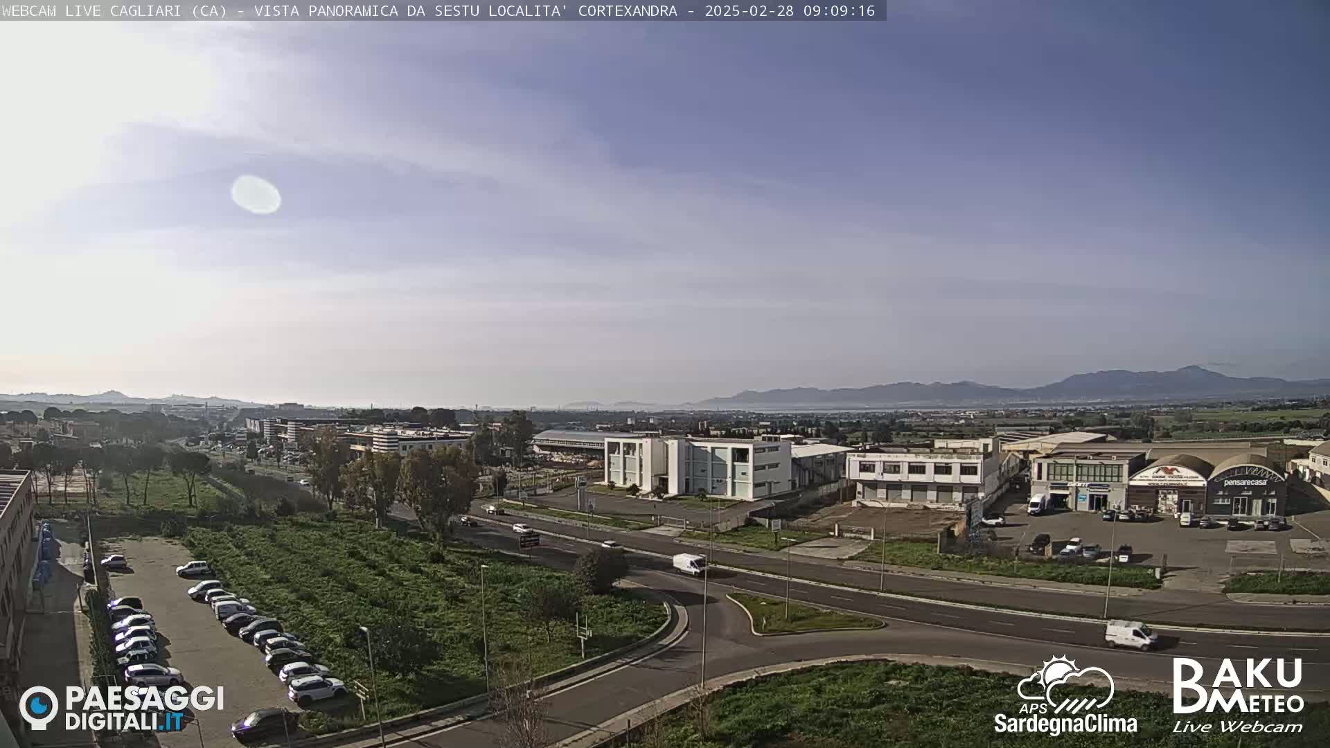 A mostly clear, bright day overlooks a sprawling industrial area with a parking lot full of cars and light traffic on roads, with distant mountains visible on the horizon.
