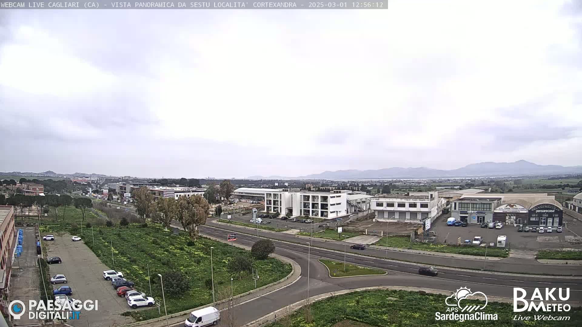 A panoramic view of a city under an overcast sky, showing roads, buildings, parked cars, and a roundabout.