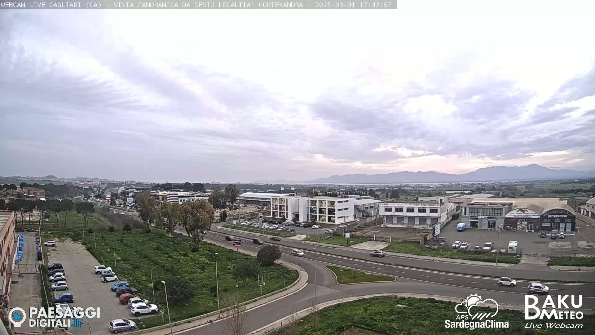 A wide, high-angle view shows a cloudy day over a suburban area with roads, buildings, and parked cars, and mountains in the distance.