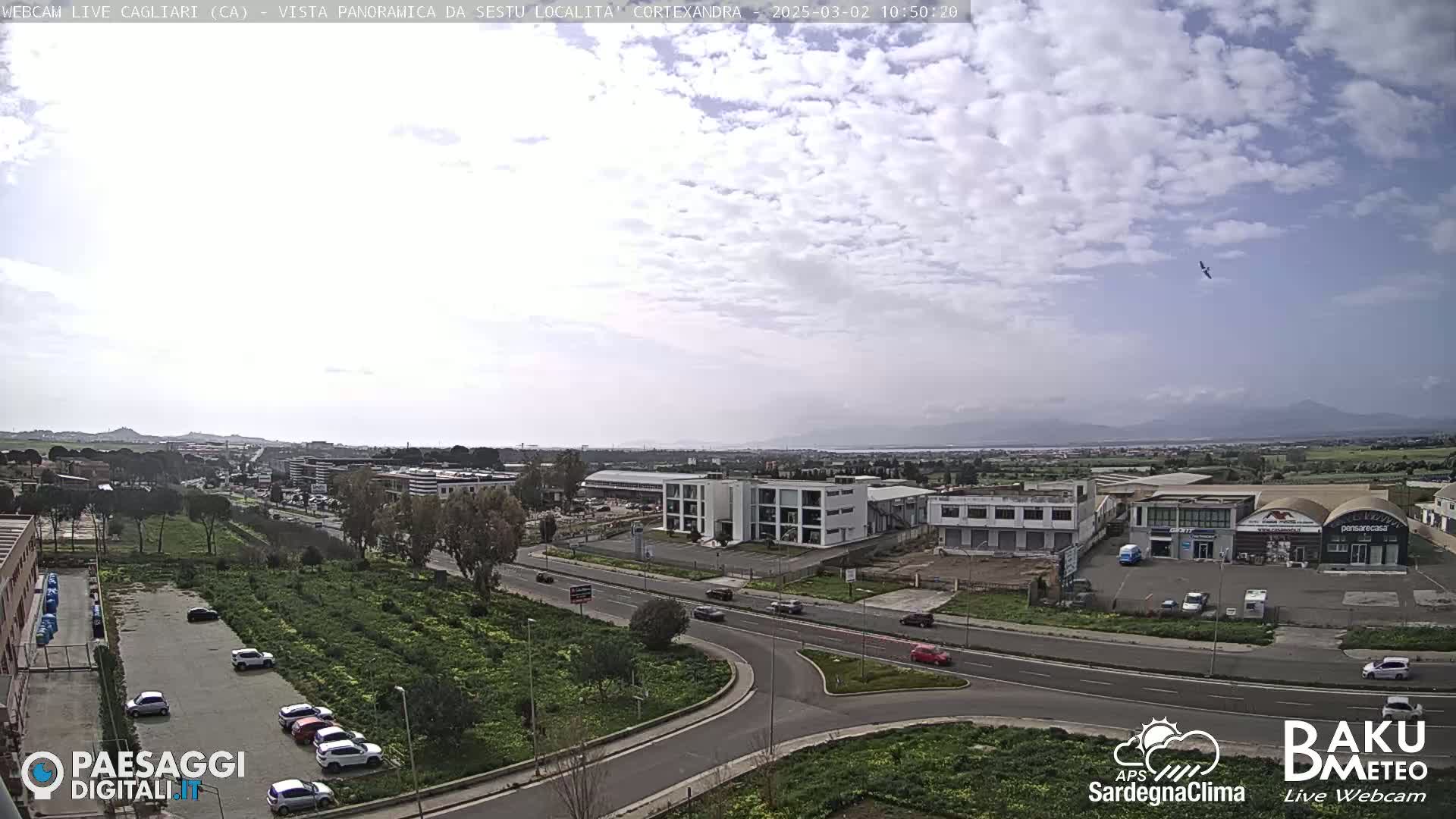 A mostly cloudy day reveals a panoramic view of a road intersection surrounded by industrial buildings and parking lots, with distant mountains and a body of water visible on the horizon.