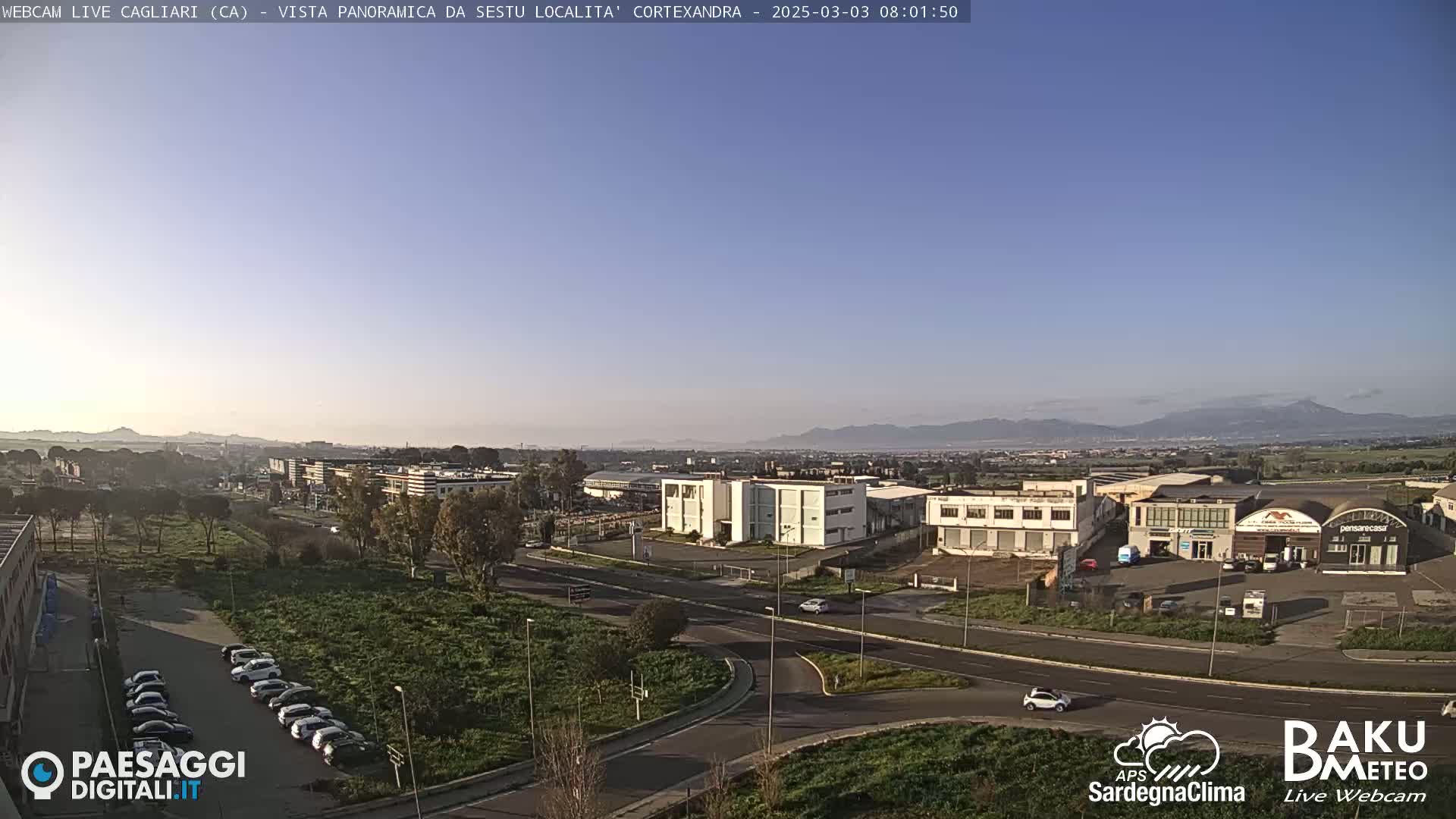 A clear, sunny day reveals a wide view of a city with industrial buildings, roads, and parked cars, mountains visible in the distance.