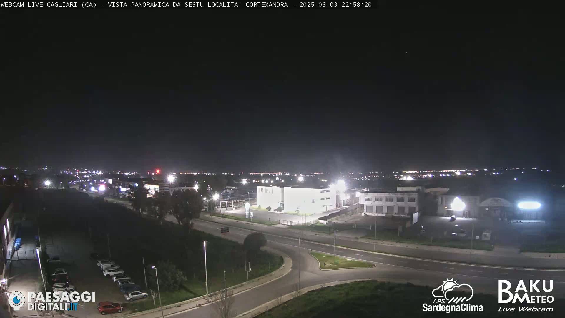 A nighttime, high-angle view of a road intersection and industrial area with a city skyline in the distance under a clear, dark sky.