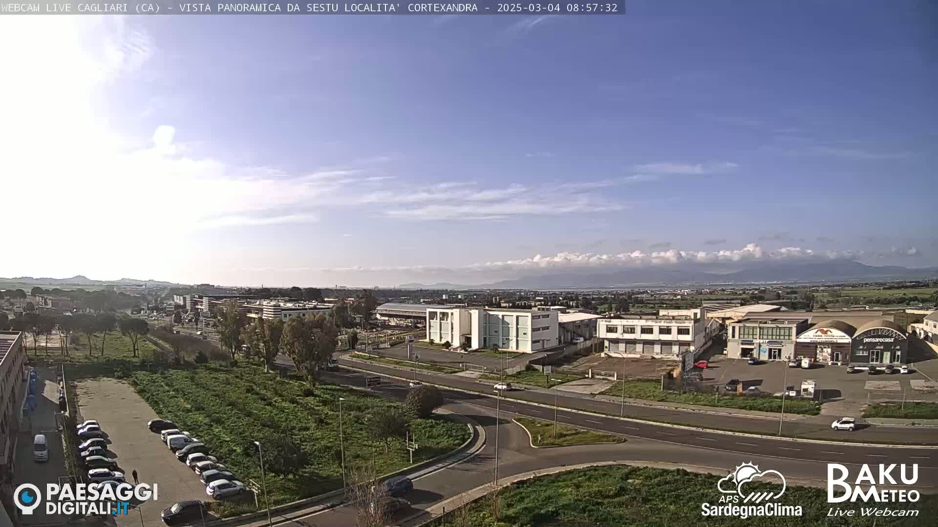 A mostly sunny day reveals a panoramic view of a city with industrial buildings, roads, and parked cars, set against a backdrop of distant mountains under a partly cloudy sky.