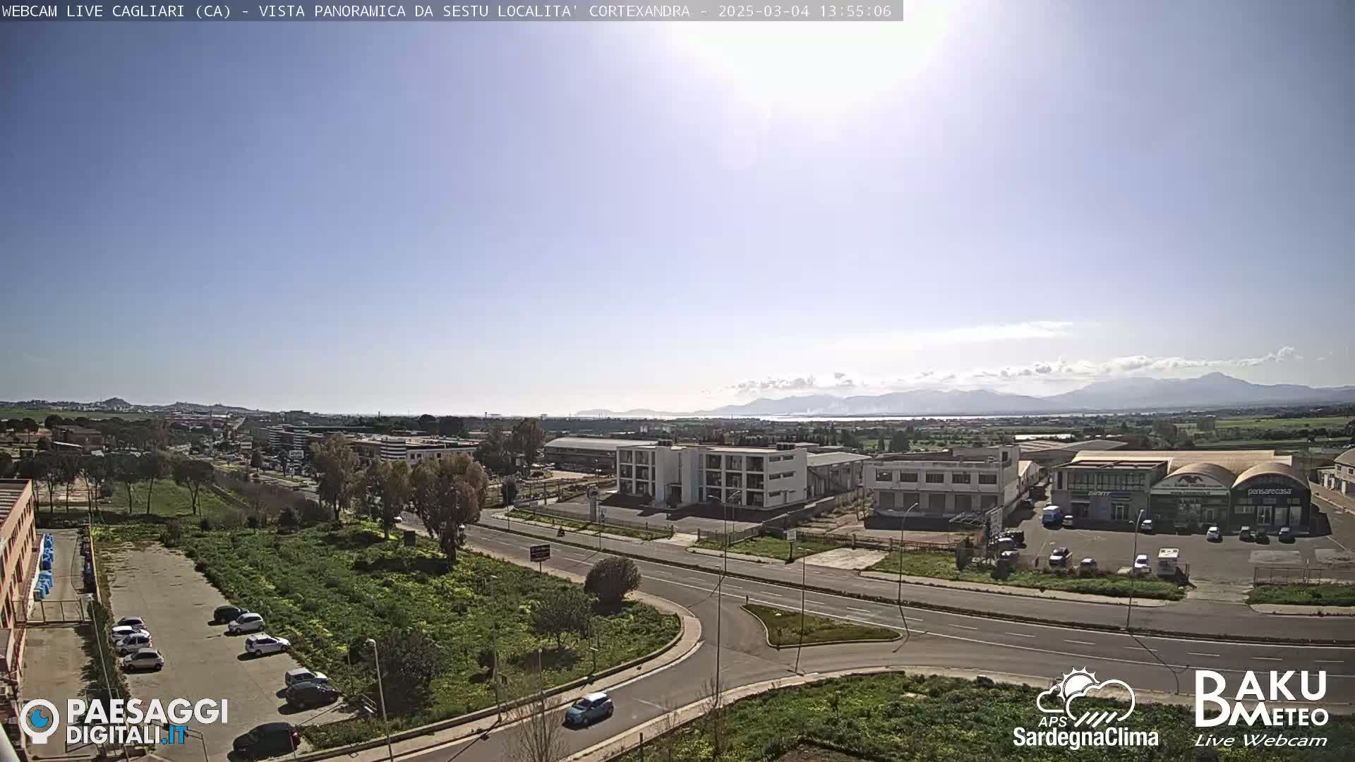 A sunny day reveals a panoramic view of a suburban area with buildings, roads, and parked cars, extending to distant mountains under a mostly clear sky.