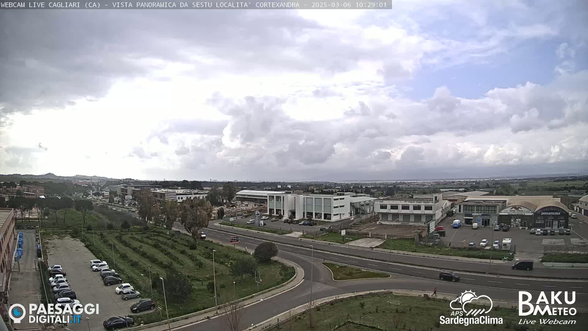 A panoramic view of a city under a partly cloudy sky shows roads, buildings, and parking lots.