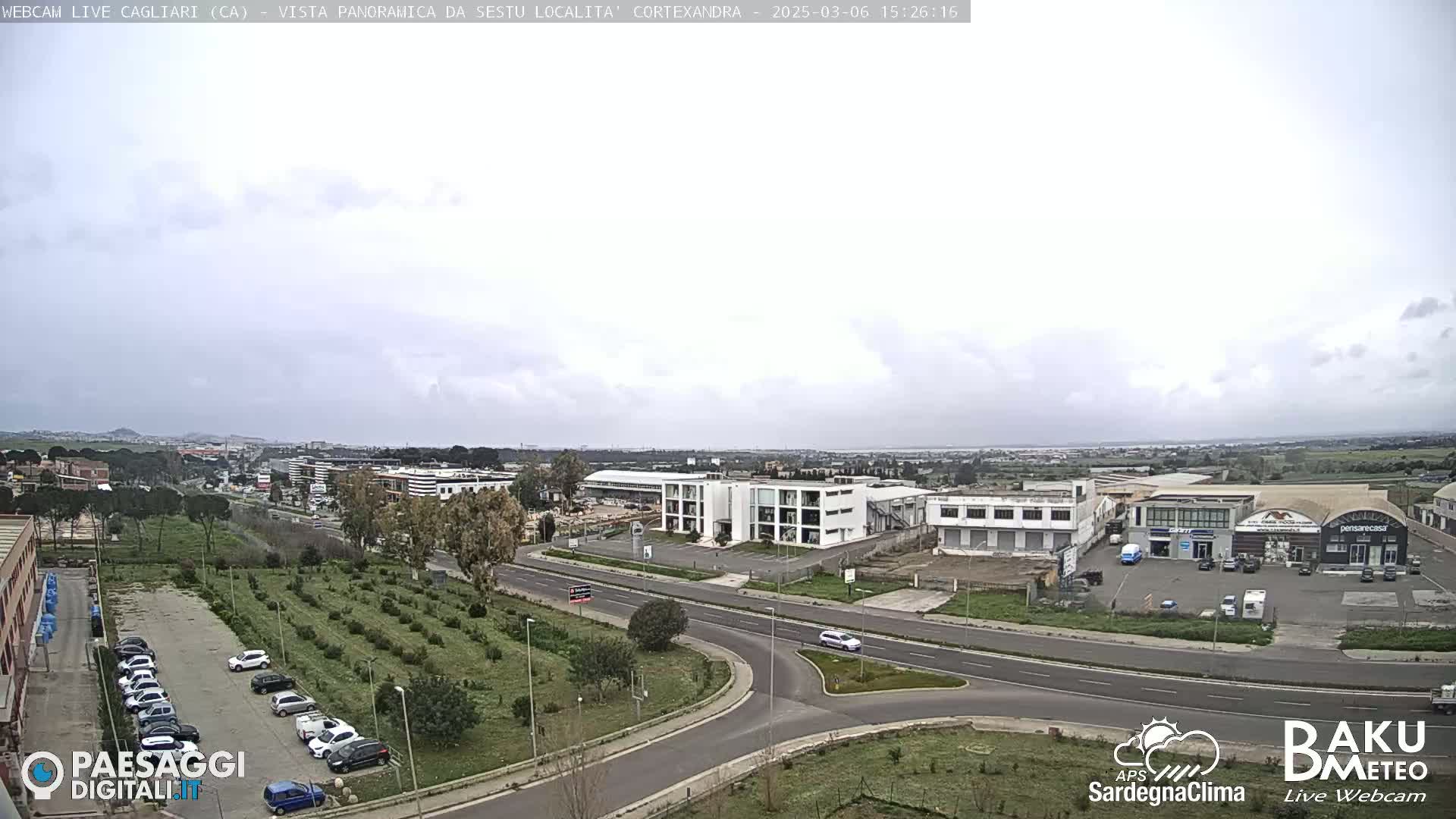 An overcast day reveals a wide aerial view of a road intersection, surrounded by industrial buildings, small businesses, and a sparse field of young trees, with cars parked in a lot and driving on the roadways.