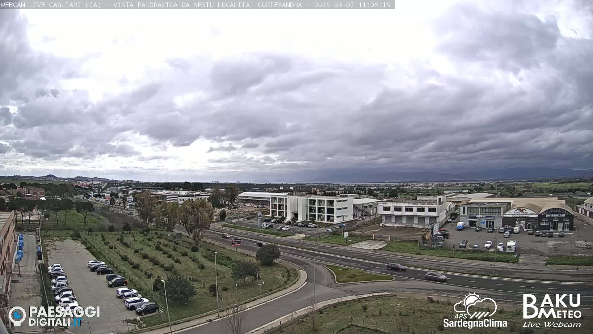 A panoramic view of a city under a cloudy sky shows roads, buildings, and parking lots filled with cars, with a large expanse of flat land in the background.