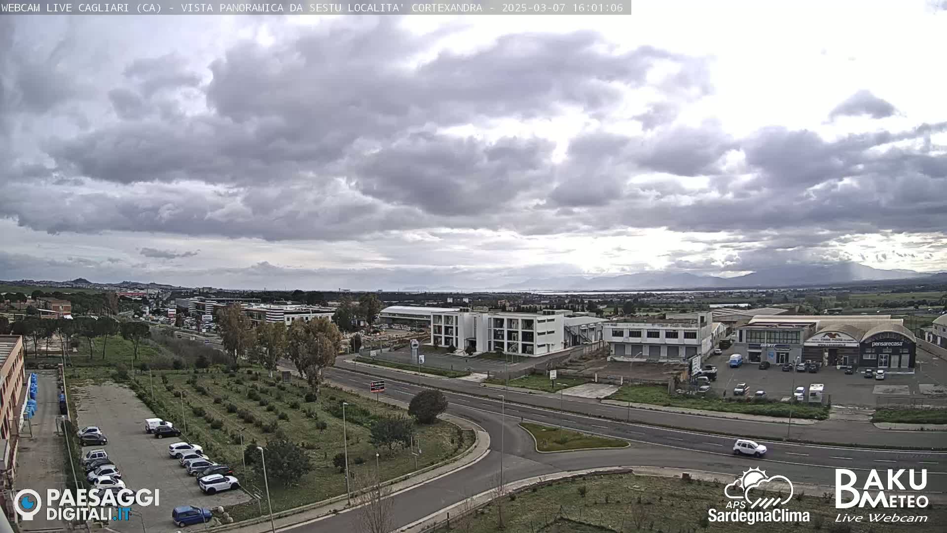 A wide, overcast view of a city landscape featuring a road intersection, buildings, and a parking lot filled with cars, with distant mountains visible on the horizon.