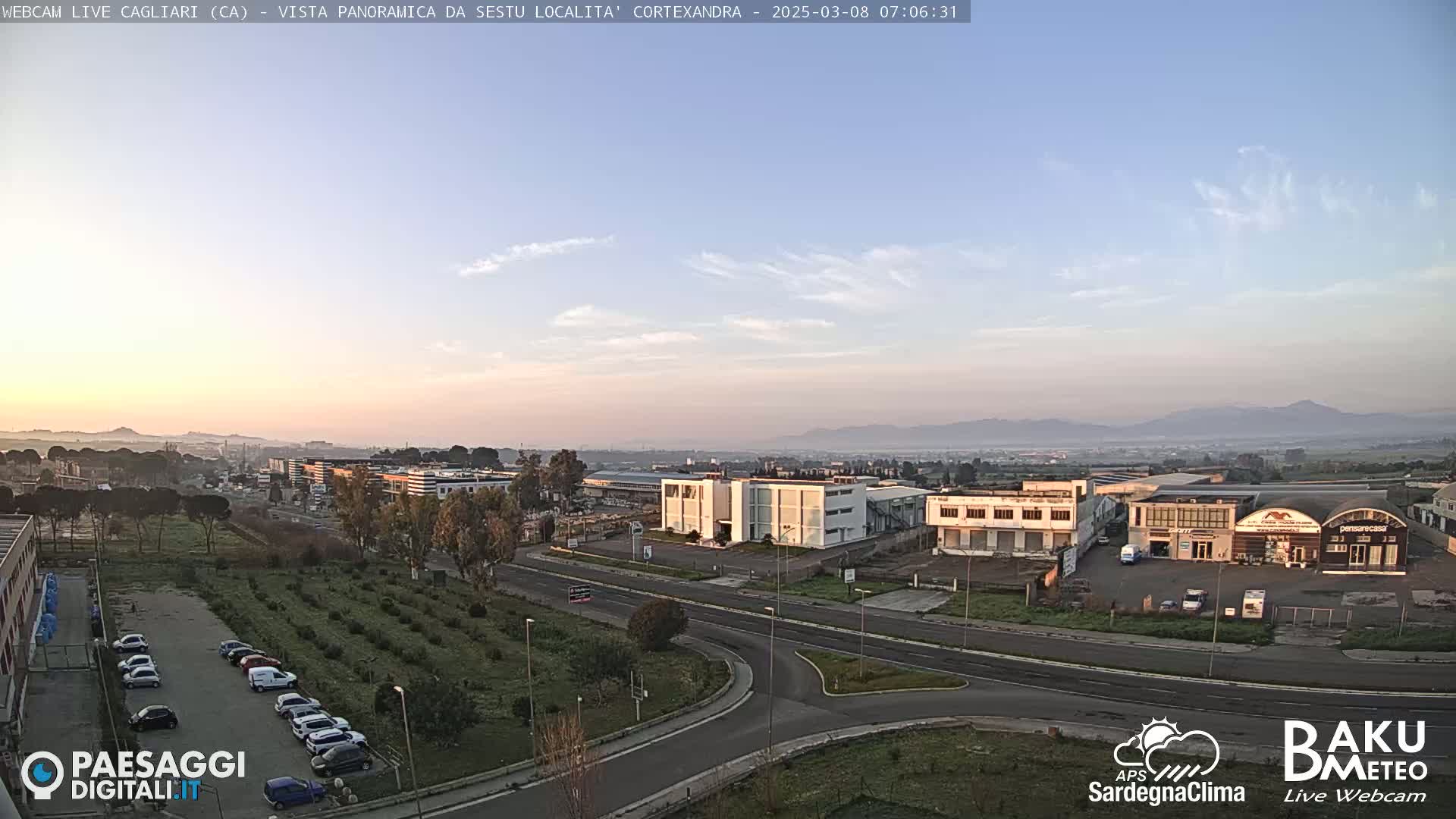 A sunrise view of an industrial area with light haze in the distance, showing buildings, roads, and a parking lot with cars.