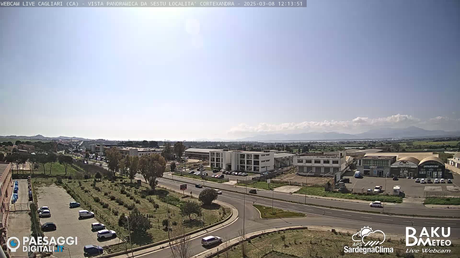 A sunny day reveals a wide shot of a roadway intersection surrounded by industrial buildings and small businesses, with a view of distant mountains and a body of water under a partly cloudy sky.