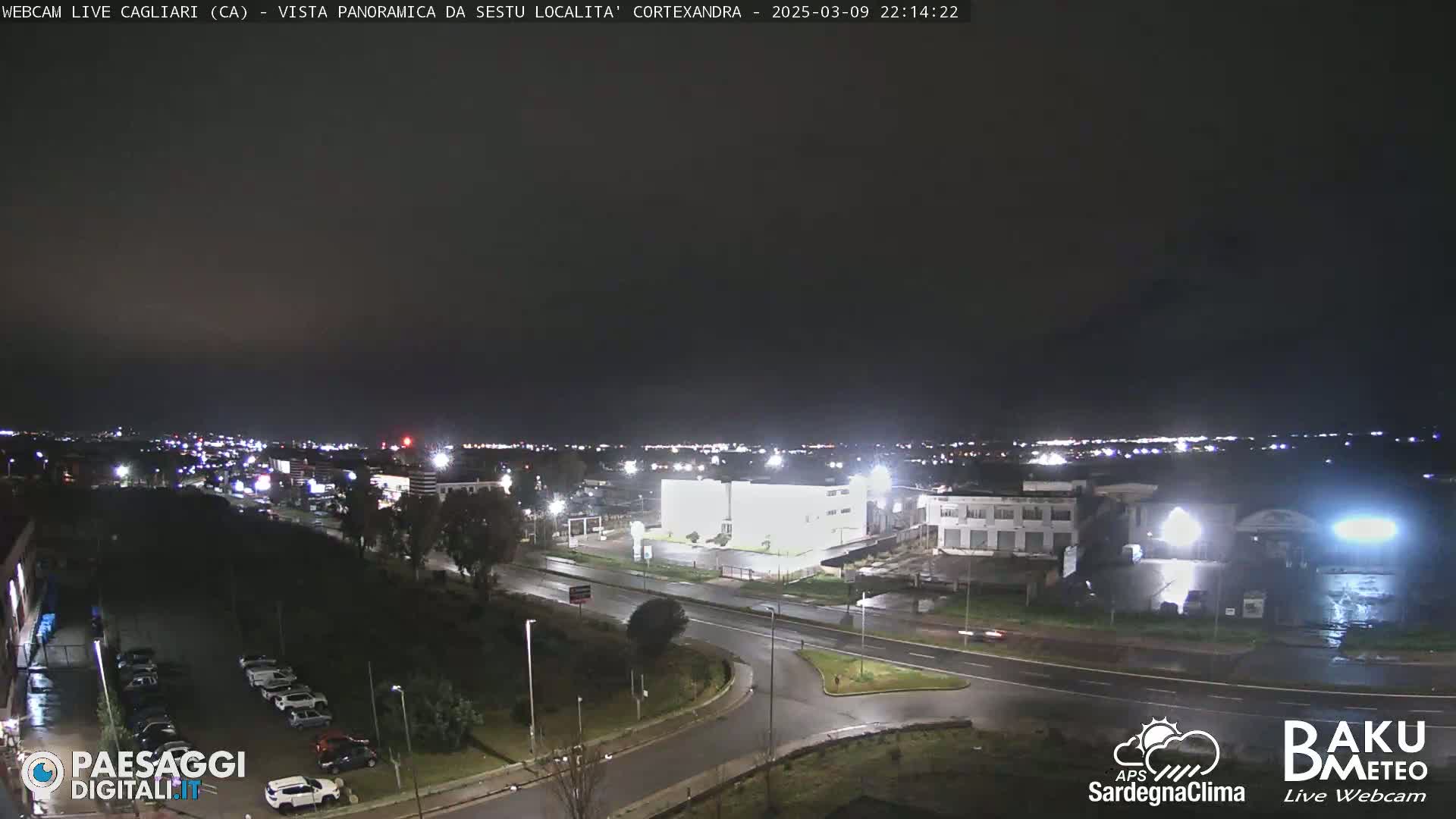 A nighttime, wet, panoramic view of a city, showing roads, buildings, and parked cars under partly cloudy skies.