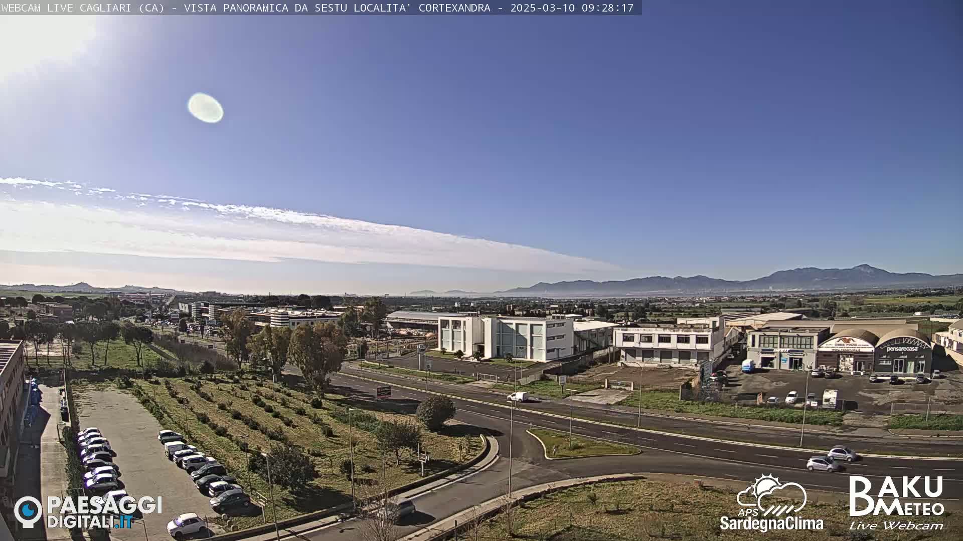 A mostly sunny day reveals a wide shot of a cityscape featuring buildings, roads, and a parking lot filled with cars, all set against a backdrop of distant mountains.