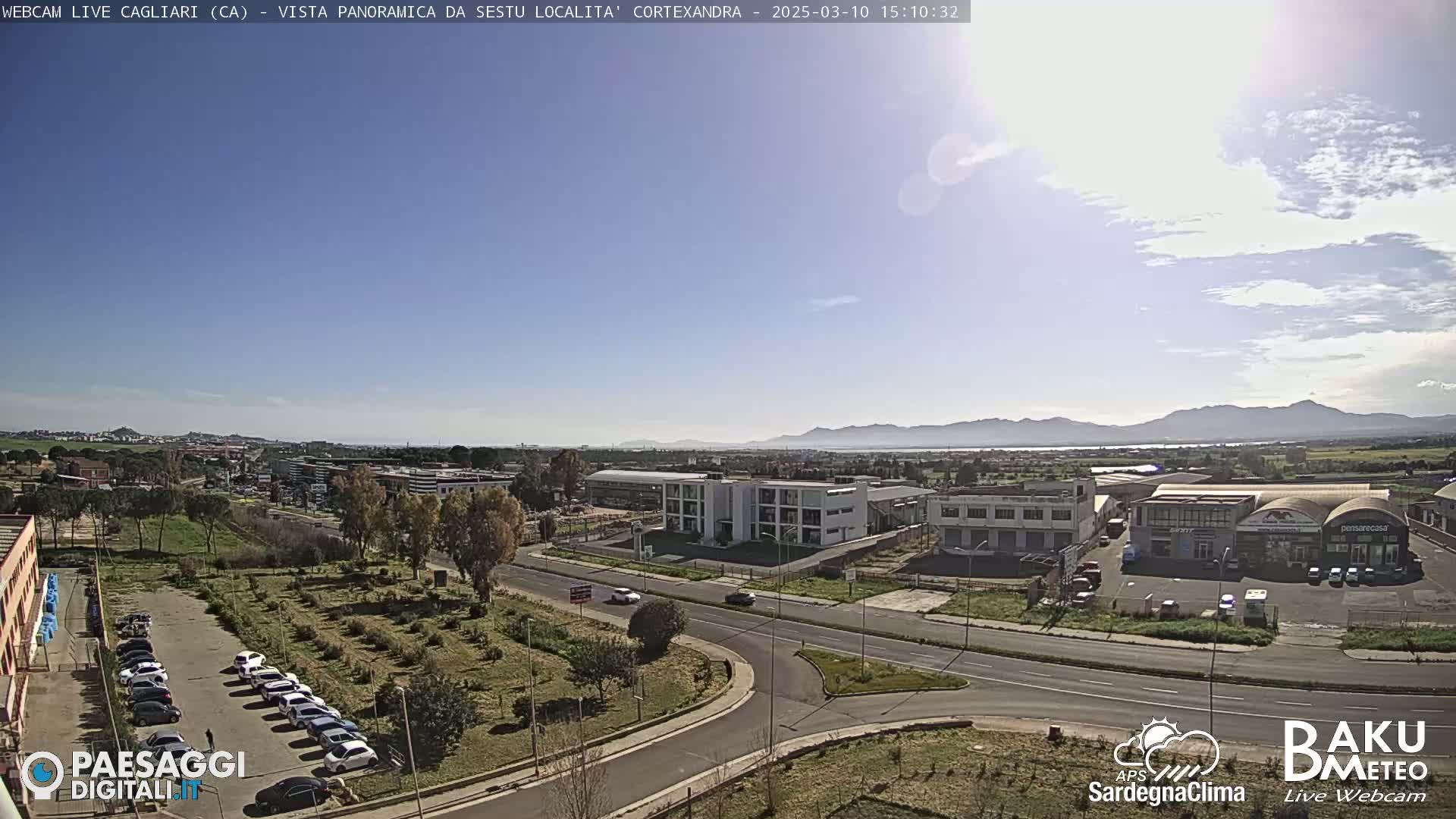 A sunny, clear day reveals a panoramic view of a city with industrial buildings, roads, and parked cars, mountains in the background.