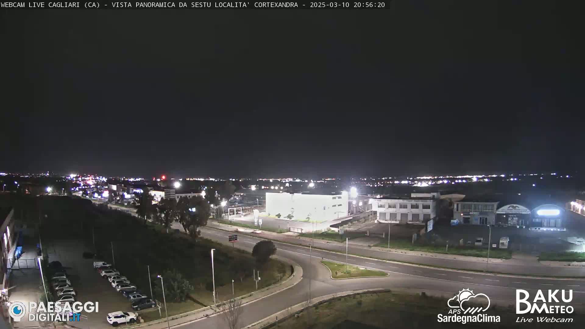 A nighttime, panoramic view of a city's outskirts, showing roads, buildings, and sparsely lit parking lots under a clear, dark sky.