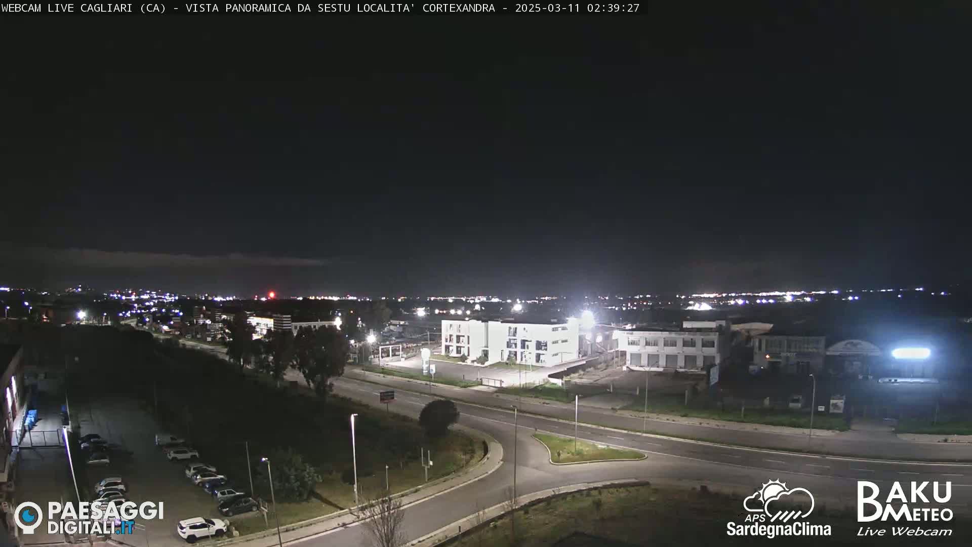 A nighttime, panoramic view of a city's outskirts, showing roads, buildings, and some parked cars under a clear, dark sky.