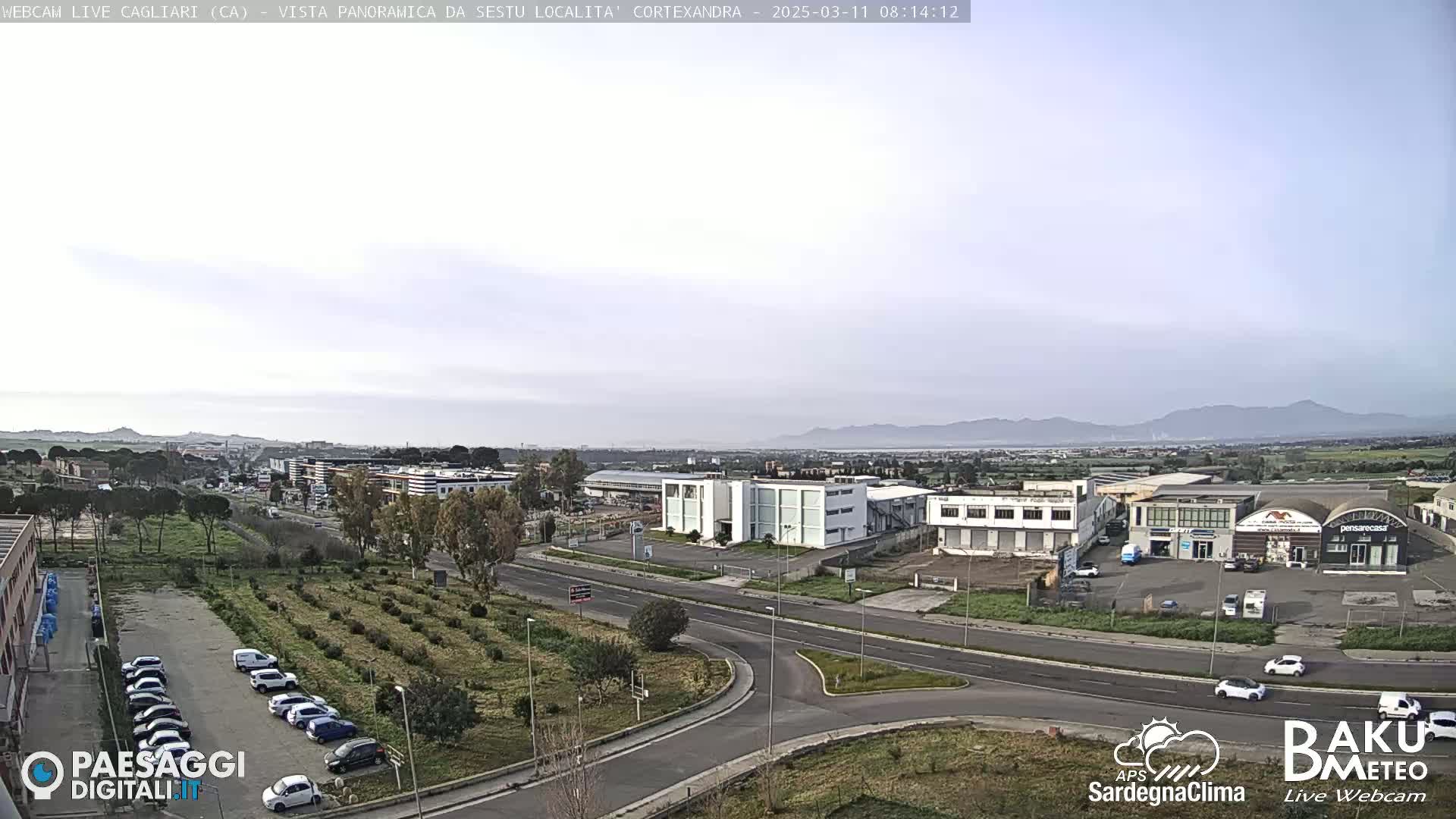 A mostly clear, daytime view of a suburban industrial area with a few cars parked in a lot, roads, and buildings, set against a backdrop of distant mountains.
