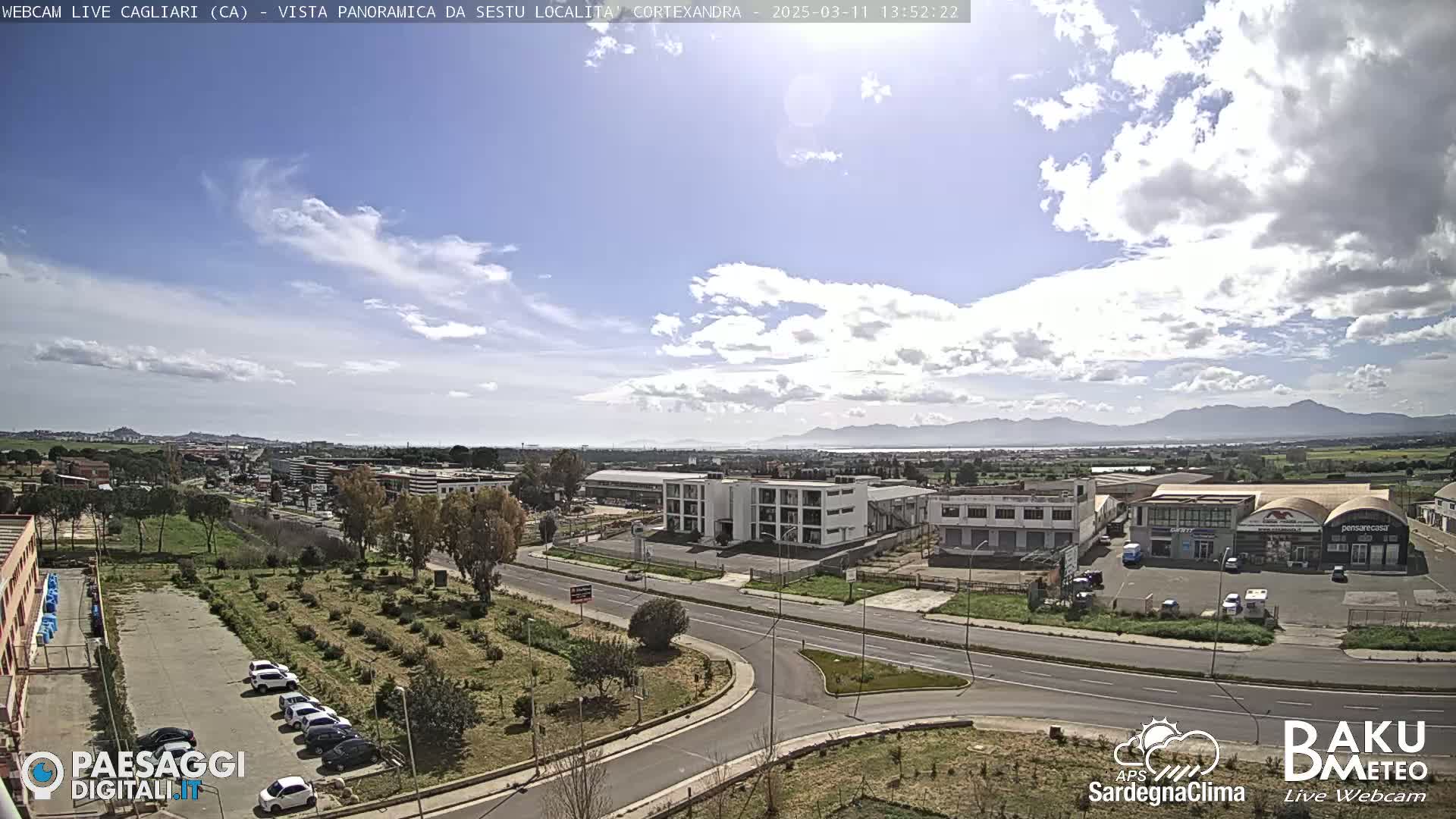 A partly cloudy day reveals a panoramic view of a suburban area with industrial buildings, roads, and parked cars, mountains visible in the distance.