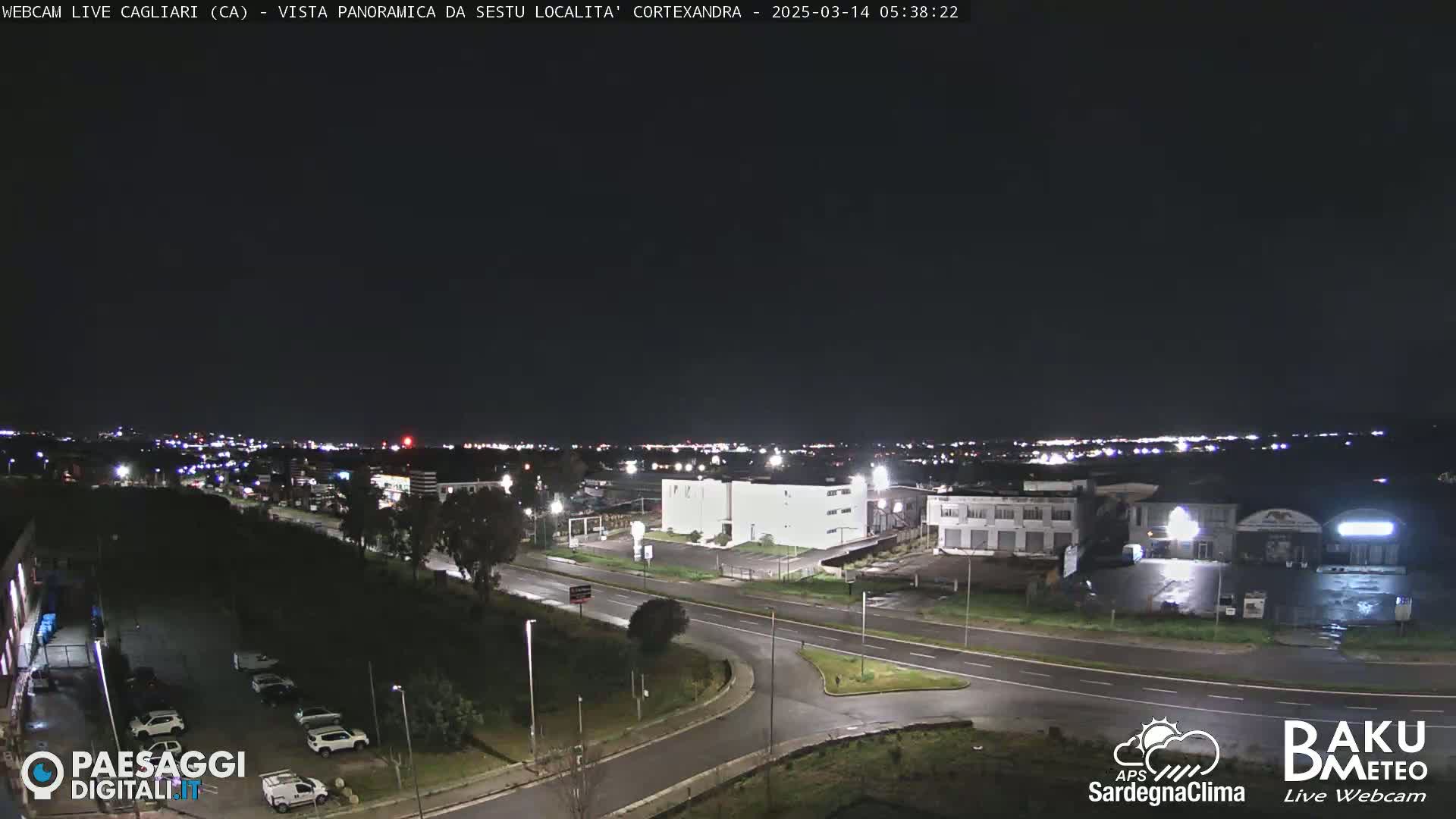 A nighttime, aerial view of a road intersection and industrial area, with a city skyline visible in the distance under a clear, dark sky.