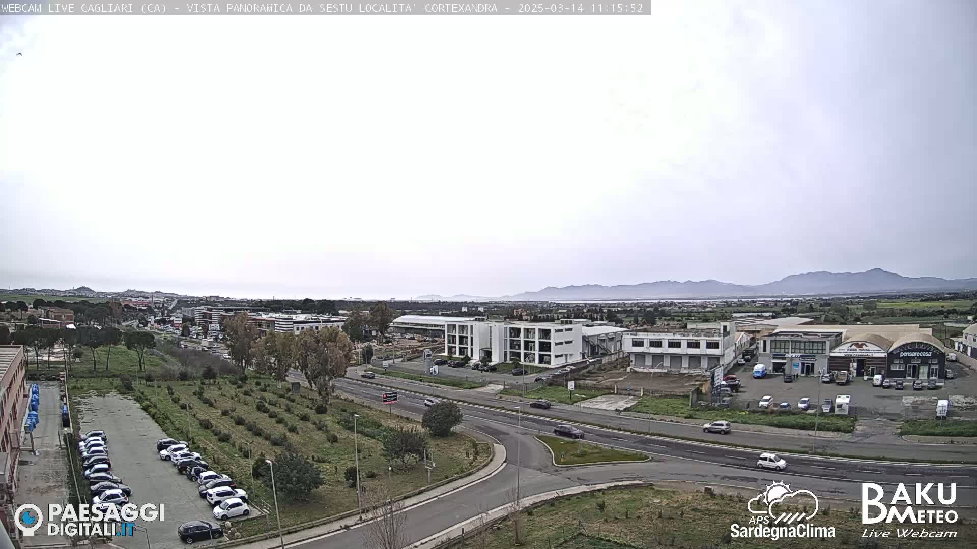 A mostly cloudy day shows a wide shot of a roadway intersection and surrounding industrial and residential areas, with distant mountains visible in the background.