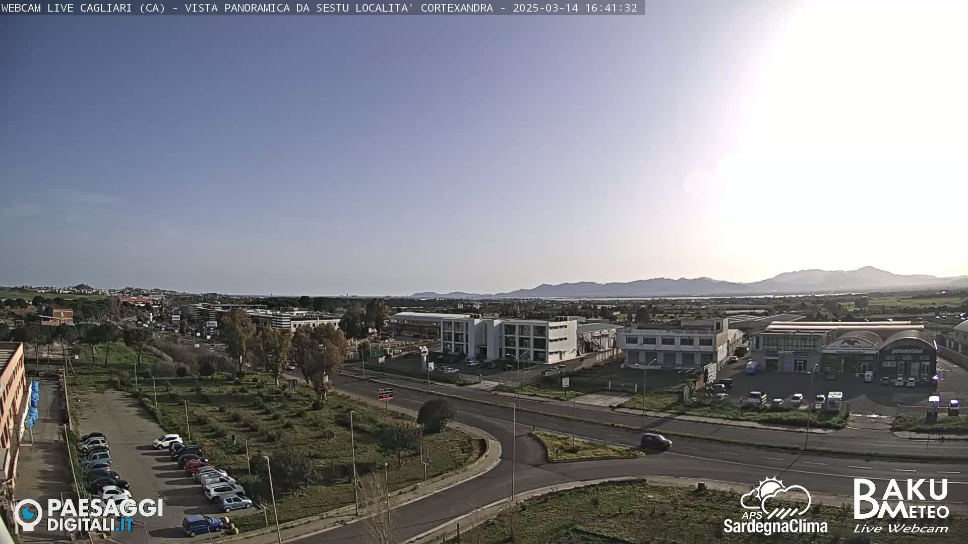 A sunny, clear day reveals a panoramic view of a city's outskirts, featuring roads, buildings, and parked cars, with distant mountains visible on the horizon.