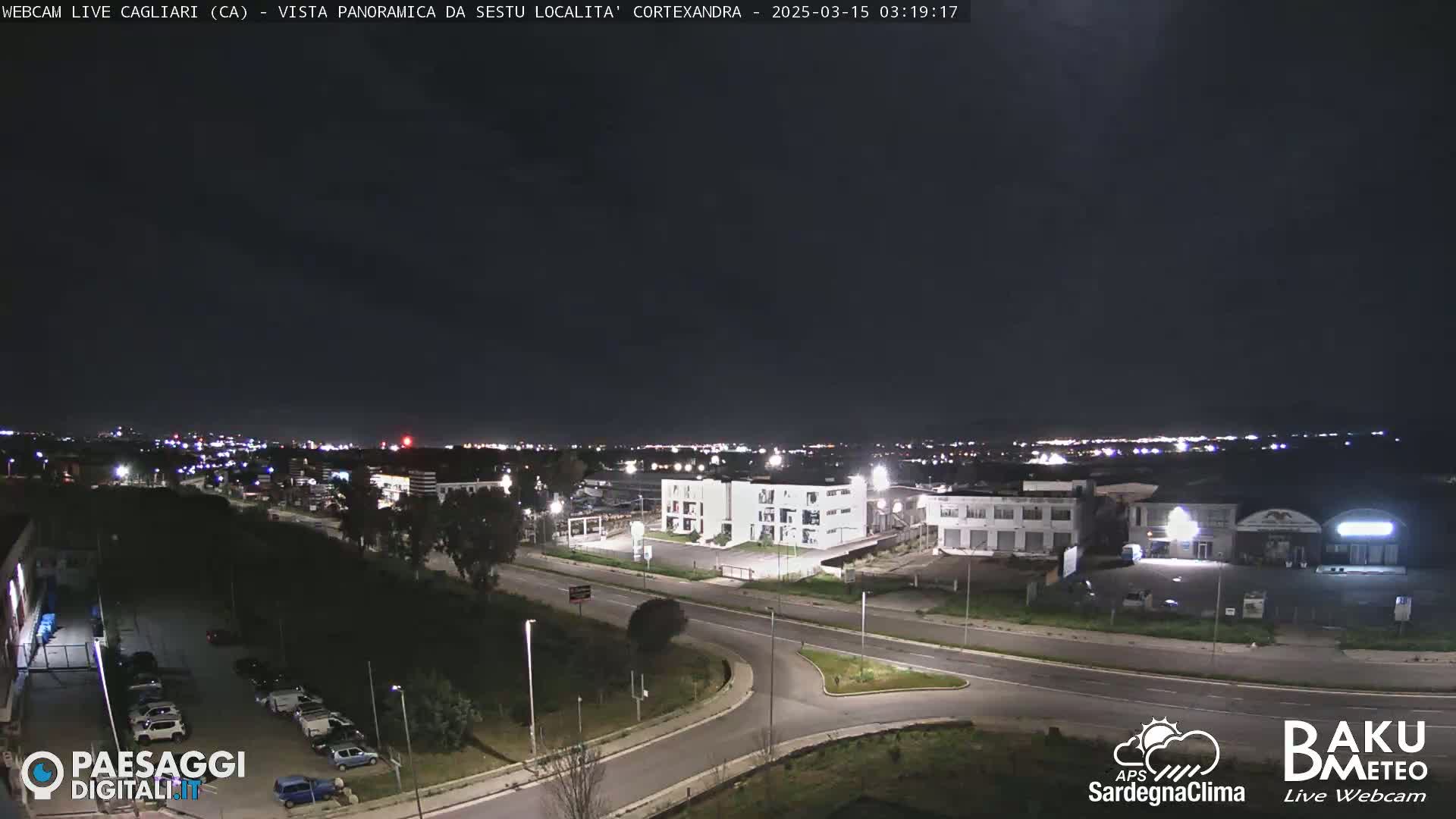 A nighttime, panoramic view of a city's outskirts, featuring a road intersection and several buildings under a clear, dark sky.