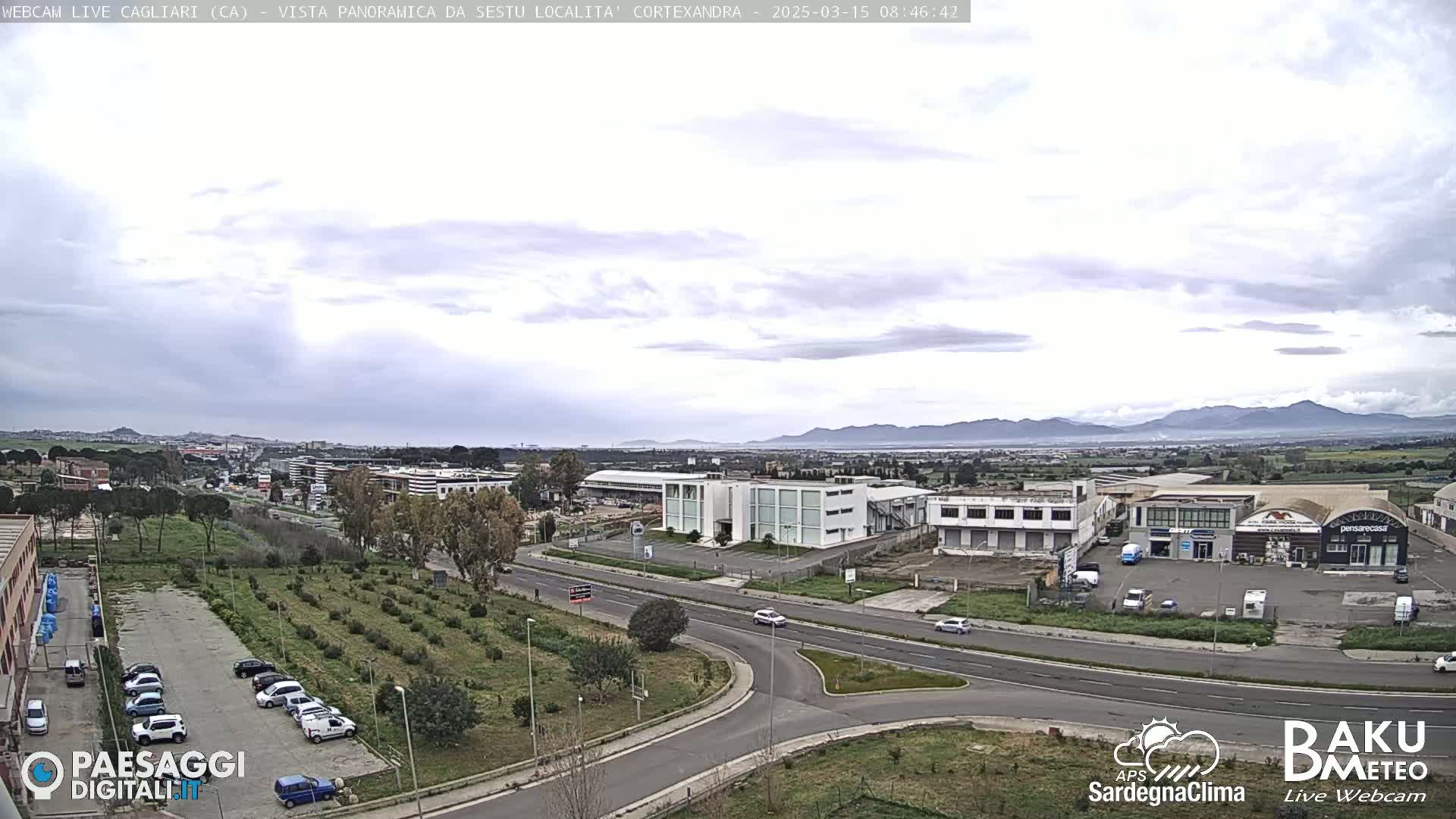 A panoramic view of a city on an overcast day shows a road intersection surrounded by buildings, parked cars, and sparsely planted trees, with distant mountains visible in the background.