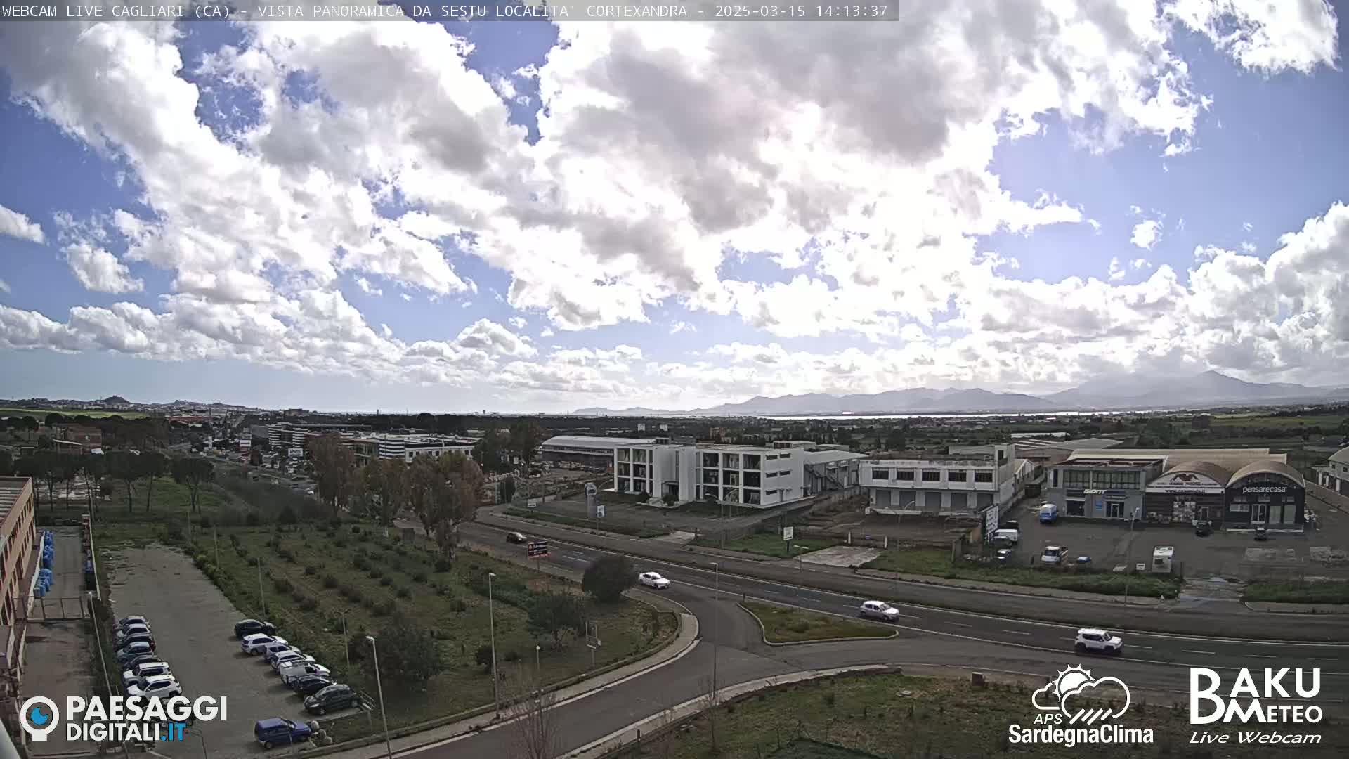 A partly cloudy day reveals a panoramic view of a city, featuring a multi-lane road, buildings, and distant mountains.