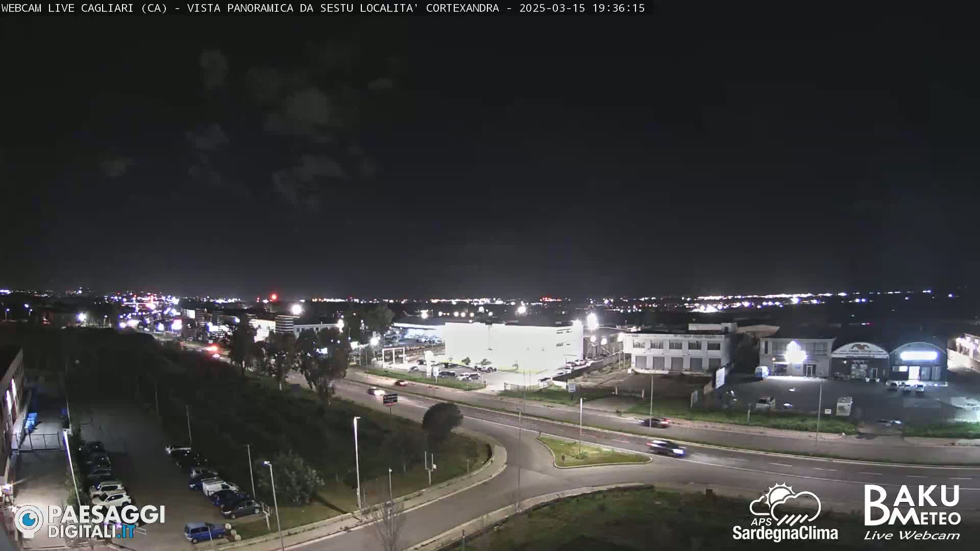 A nighttime, panoramic view of a city's outskirts, featuring roadways, buildings, and sparse vegetation under a mostly clear sky.
