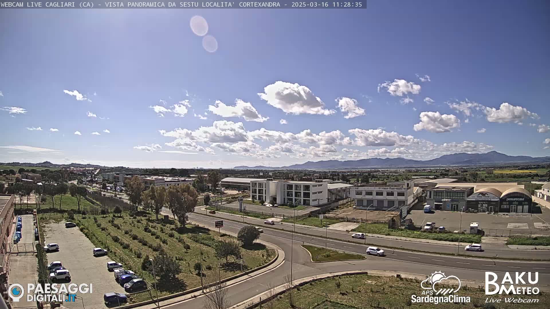 A partly cloudy day reveals a wide view of a city's outskirts, featuring roads, buildings, and a parking lot, with mountains visible in the distance.