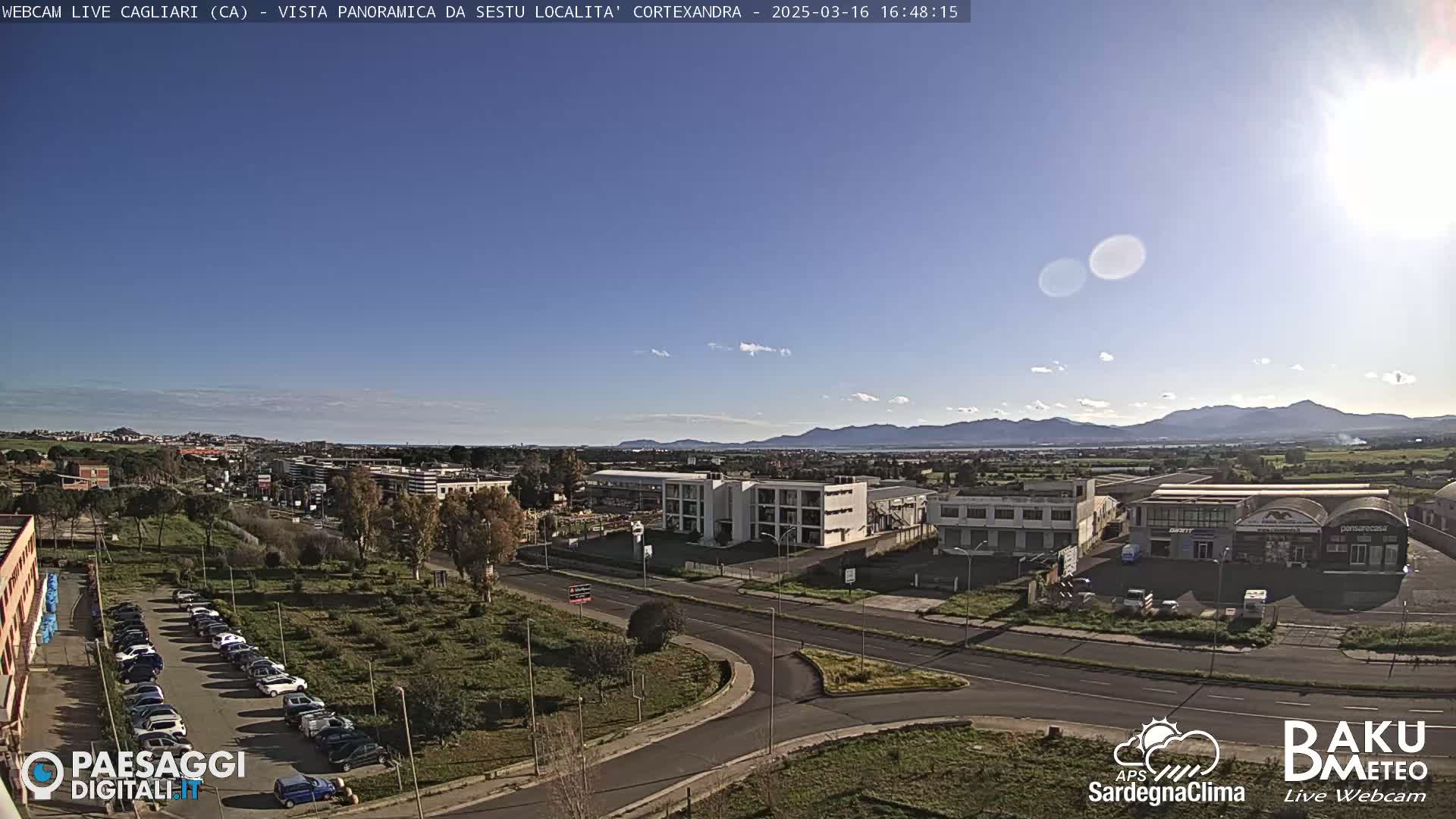 A sunny day reveals a panoramic view of a city with industrial buildings, a parking lot, and a roadway, all set against a backdrop of distant mountains under a clear blue sky.
