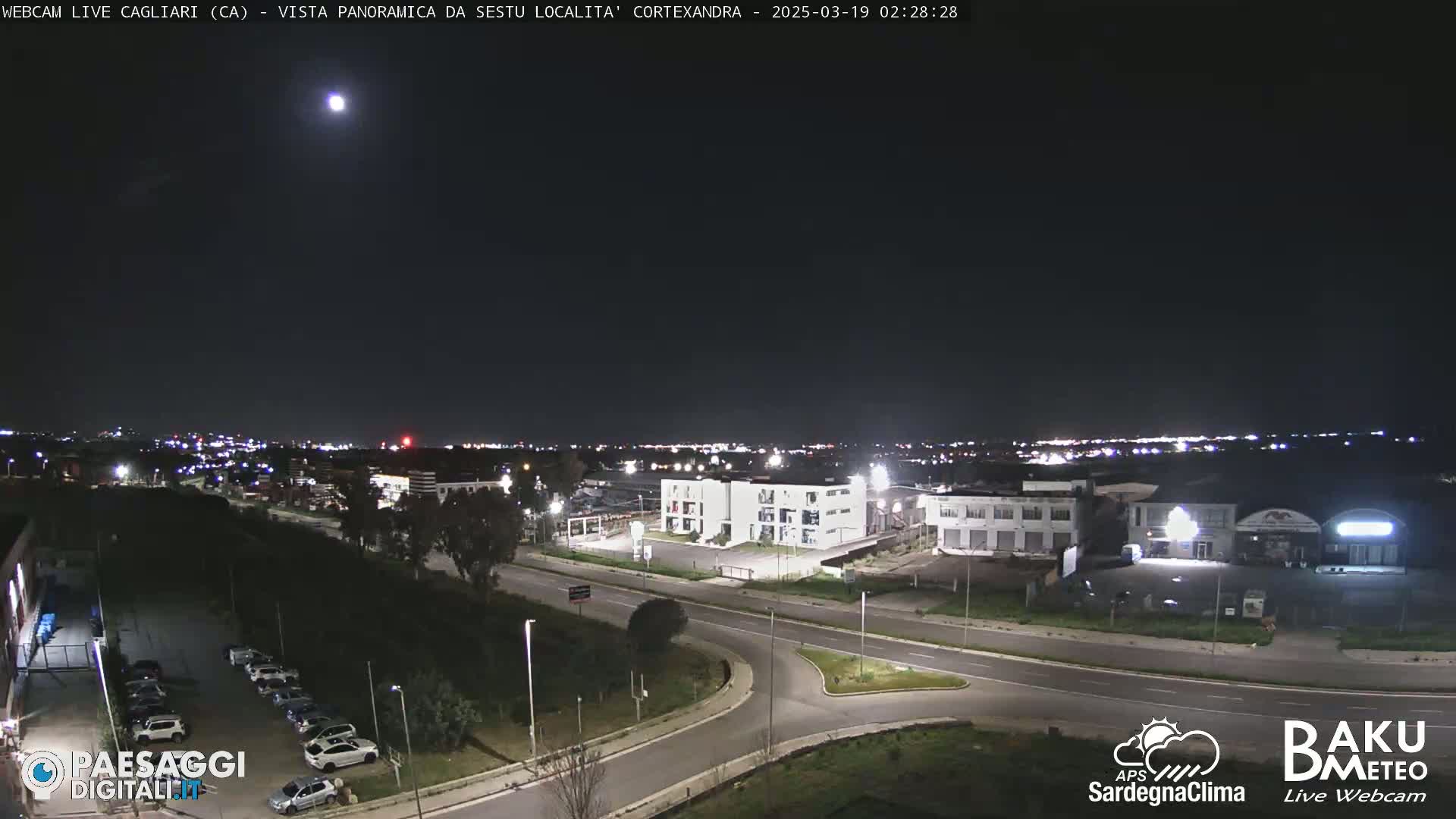 A nighttime, panoramic view of a city’s outskirts, showing roads, buildings, and parked cars under a clear, moonlit sky.
