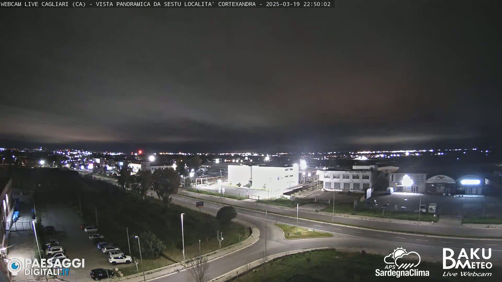 A nighttime, overcast view of a city intersection and surrounding buildings, with a parking lot and distant city lights visible.
