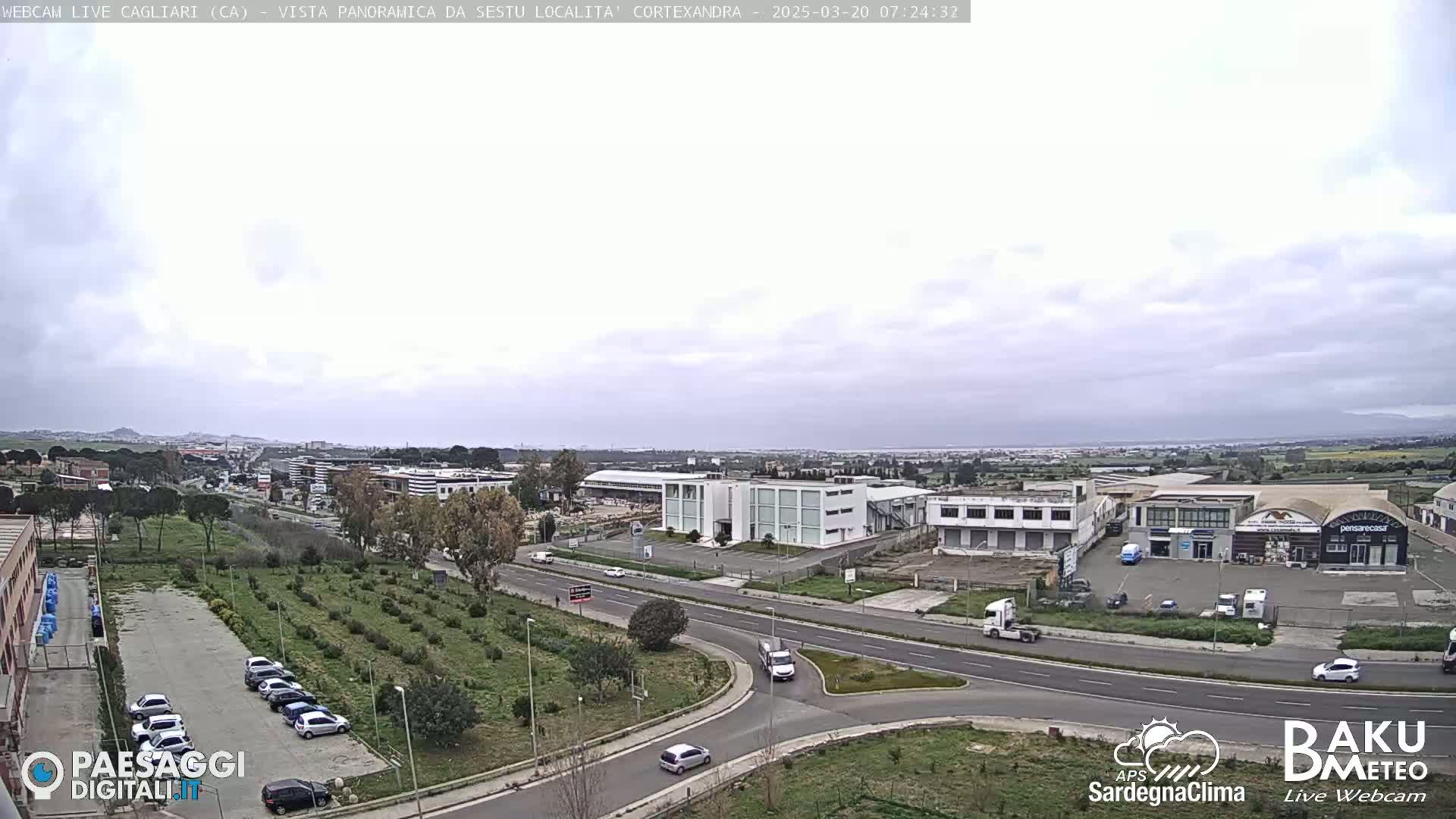 A mostly cloudy day reveals a panoramic view of an industrial area and surrounding landscape, with a road and some parked cars visible.