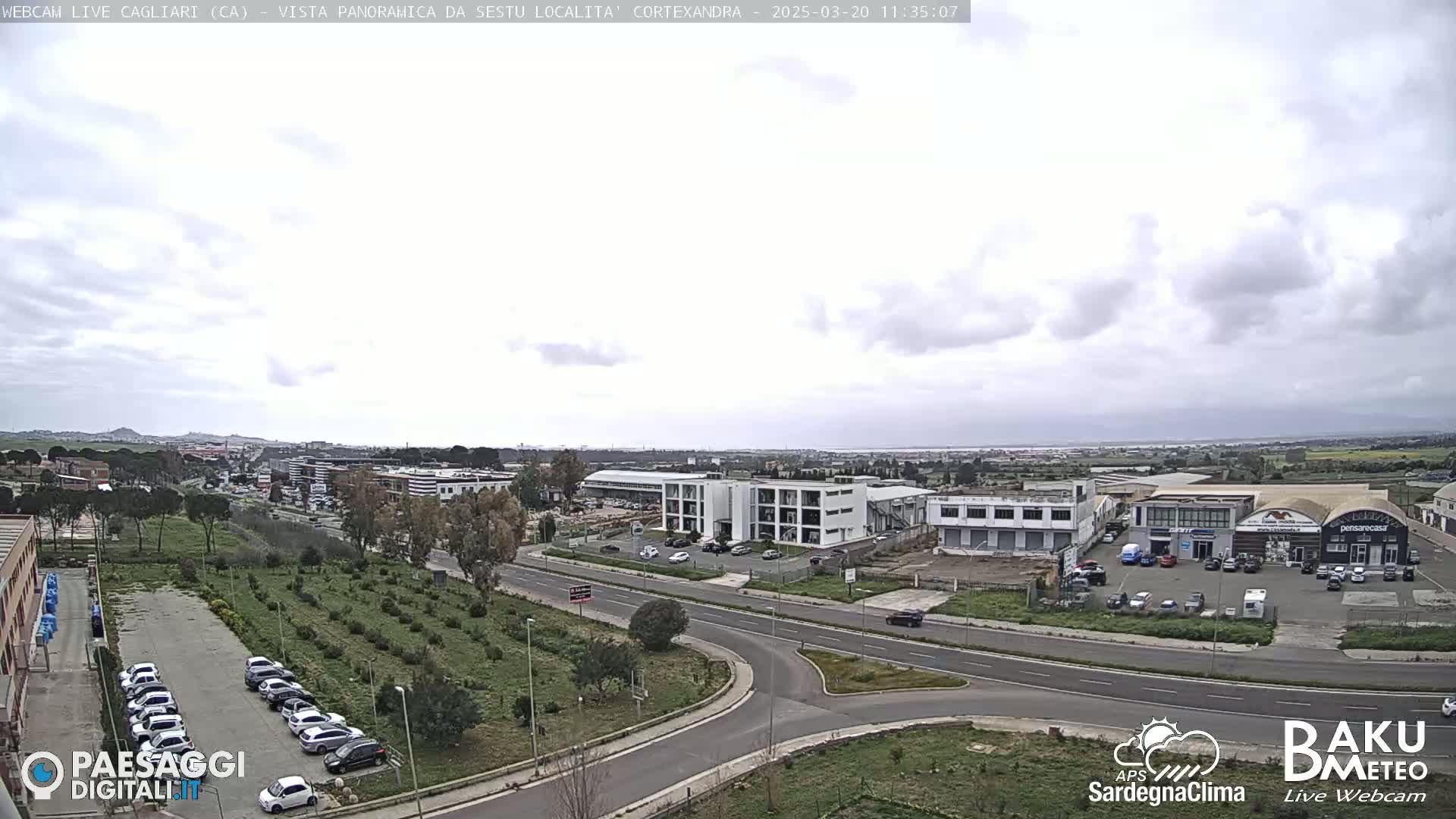 A mostly cloudy day reveals a panoramic view of a city's outskirts, showcasing roads, buildings, parked cars, and a sparsely vegetated area.