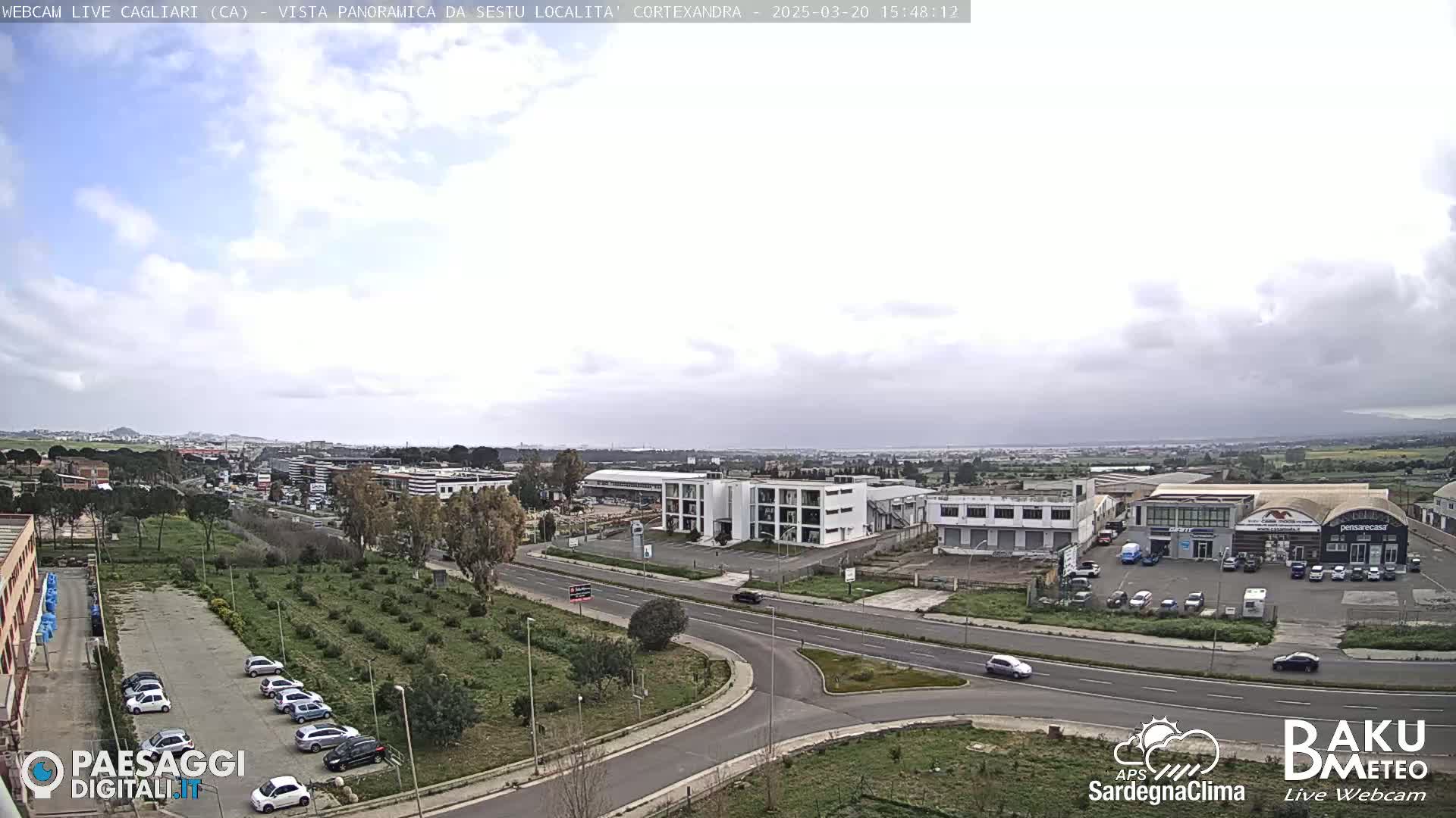 A partly cloudy day reveals a panoramic view of a city, featuring roads, buildings, and a parking lot with cars, interspersed with green areas.