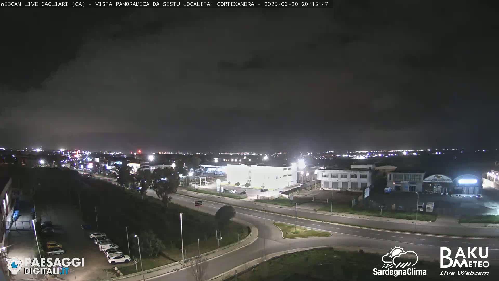 A nighttime, overcast view of a road intersection and surrounding buildings and parking lots, with city lights visible in the distance.