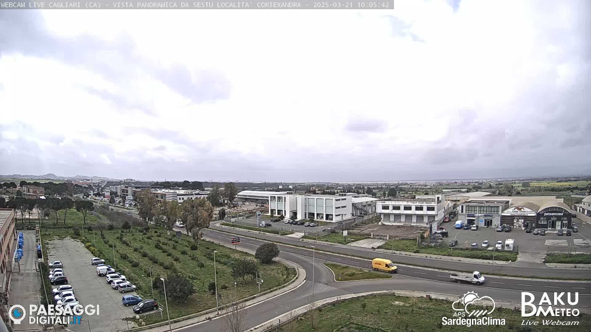 A mostly cloudy day reveals a panoramic view of a suburban area with roads, buildings, and a parking lot filled with cars.