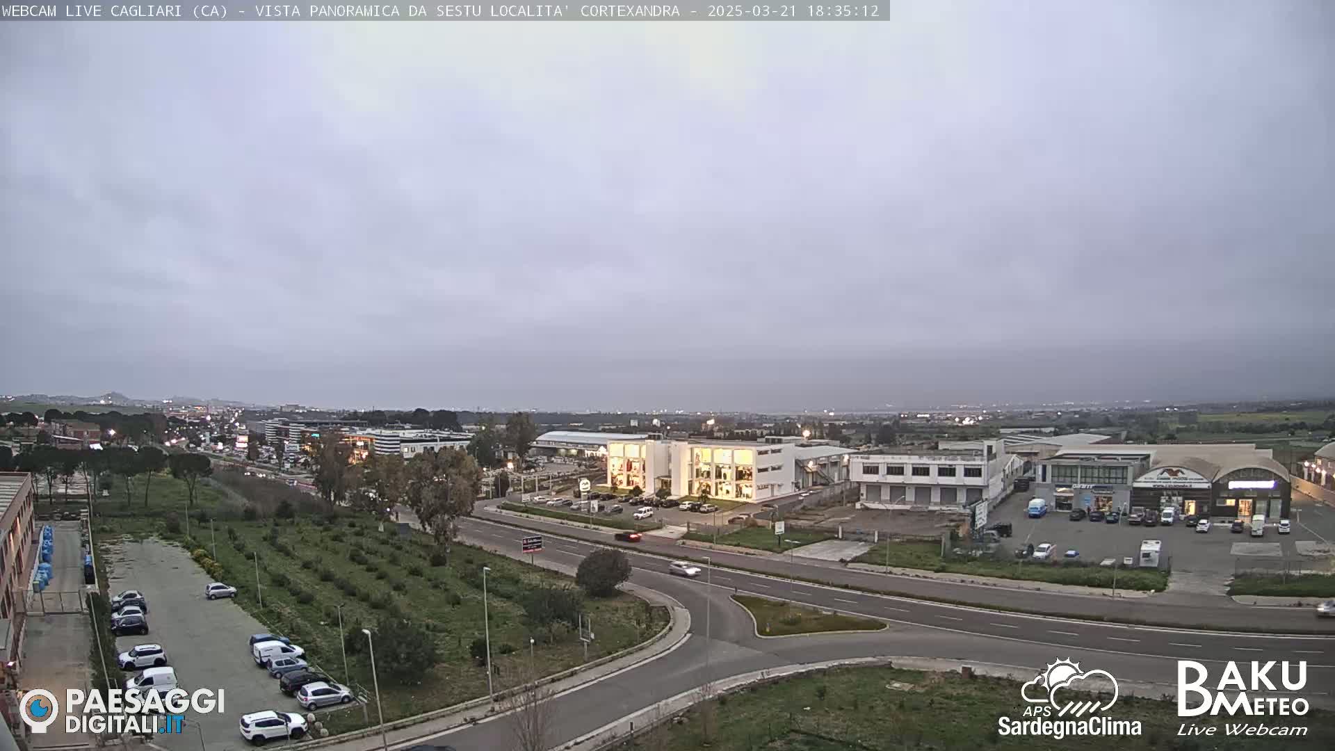 A panoramic view of a road intersection near industrial buildings and a parking lot under an overcast sky at dusk.