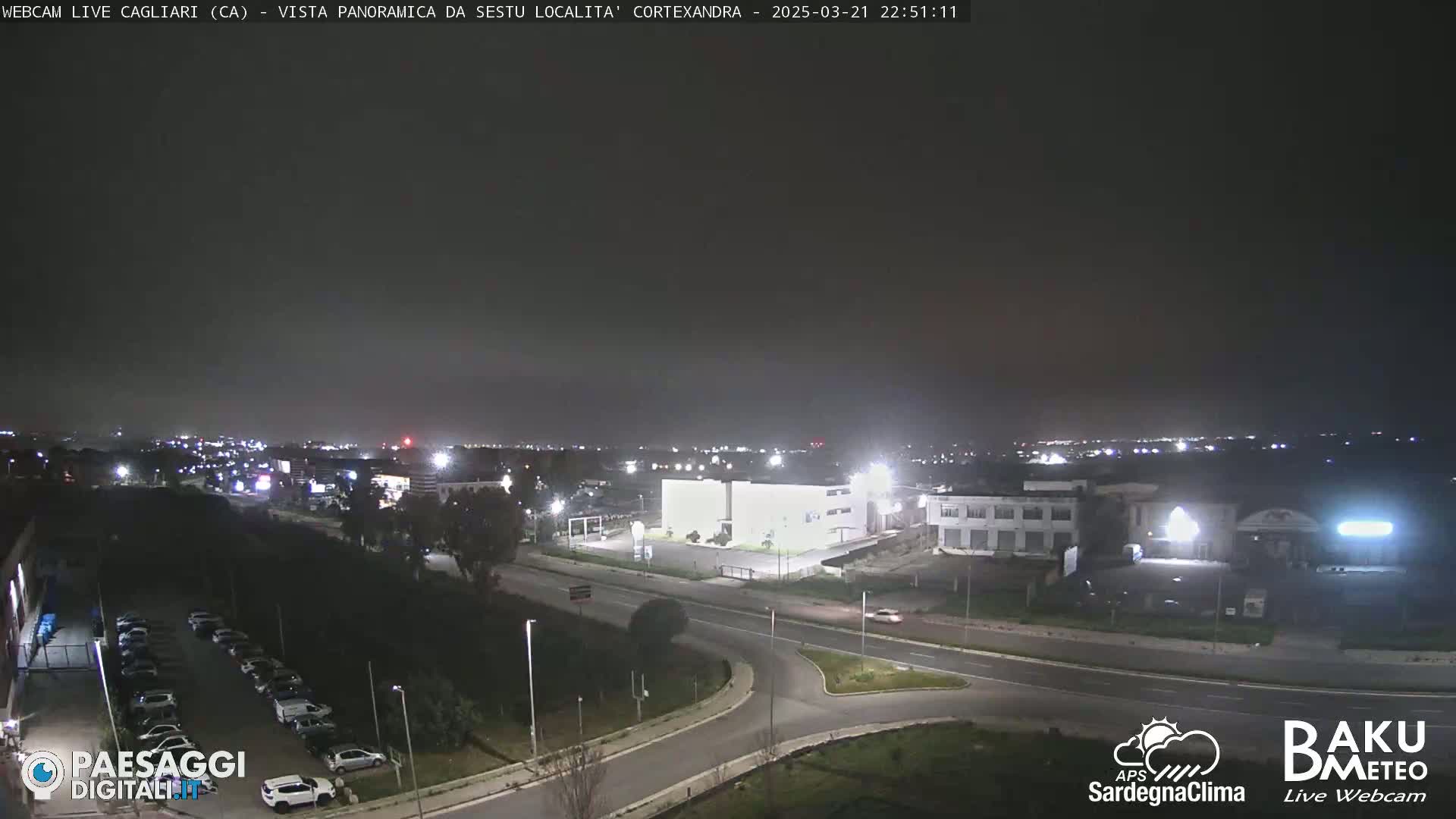 A nighttime, slightly hazy view of a city intersection and surrounding buildings, with cars parked in a lot and a few driving on the road.