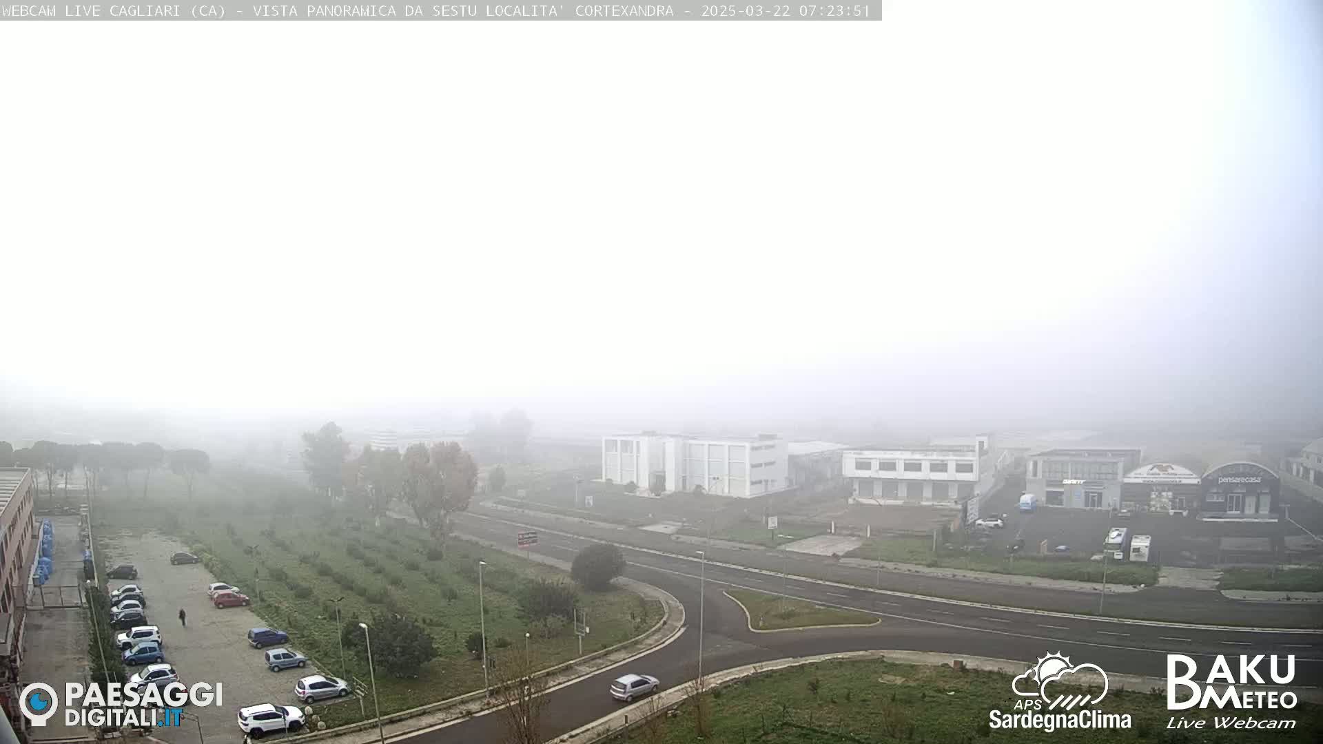 A foggy view of a road intersection and industrial buildings, with several cars parked in lots and a few driving on the road.