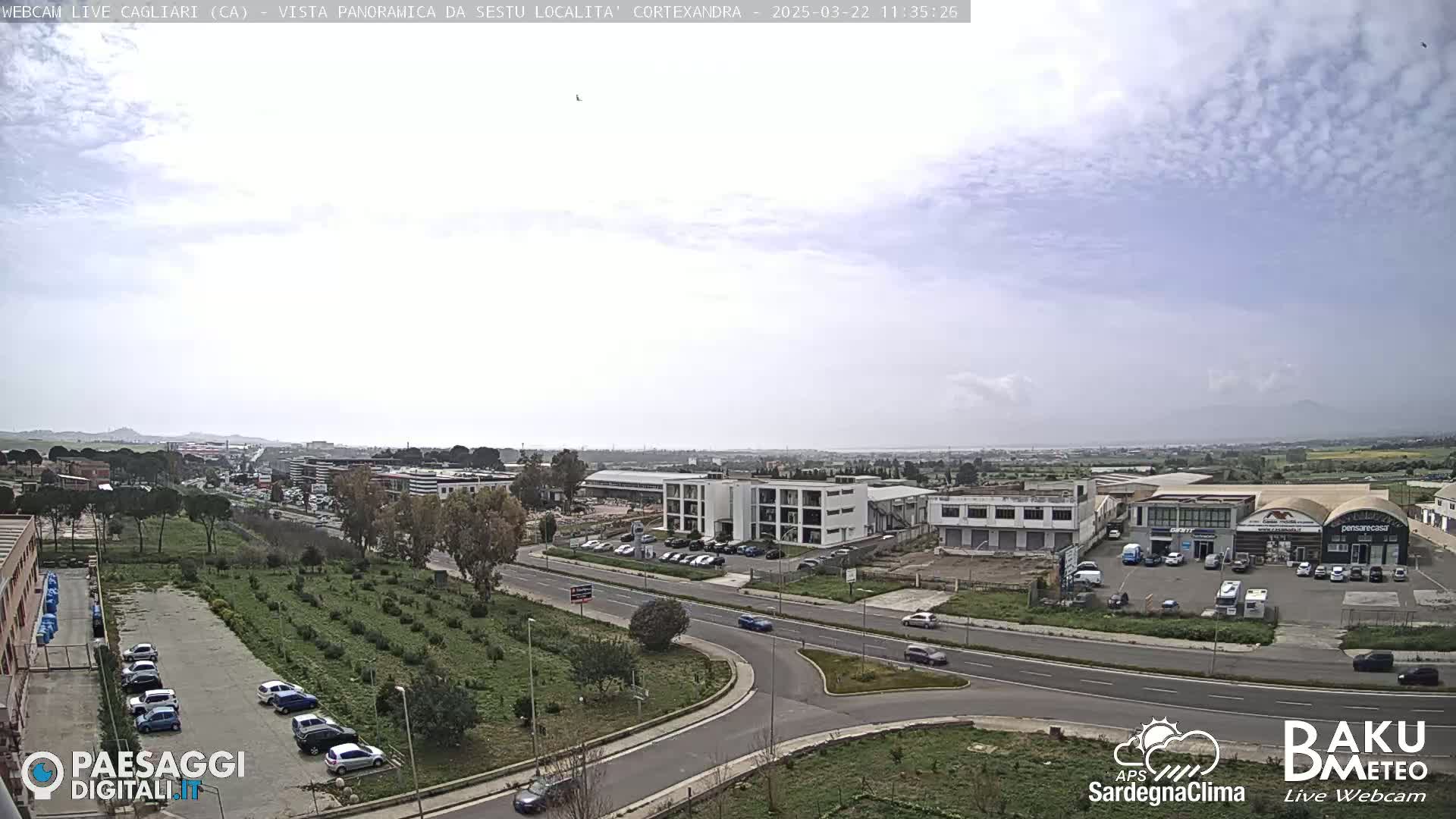 A mostly cloudy day reveals a panoramic view of a suburban area with roads, buildings, and a parking lot filled with cars, adjacent to a field of small plants.