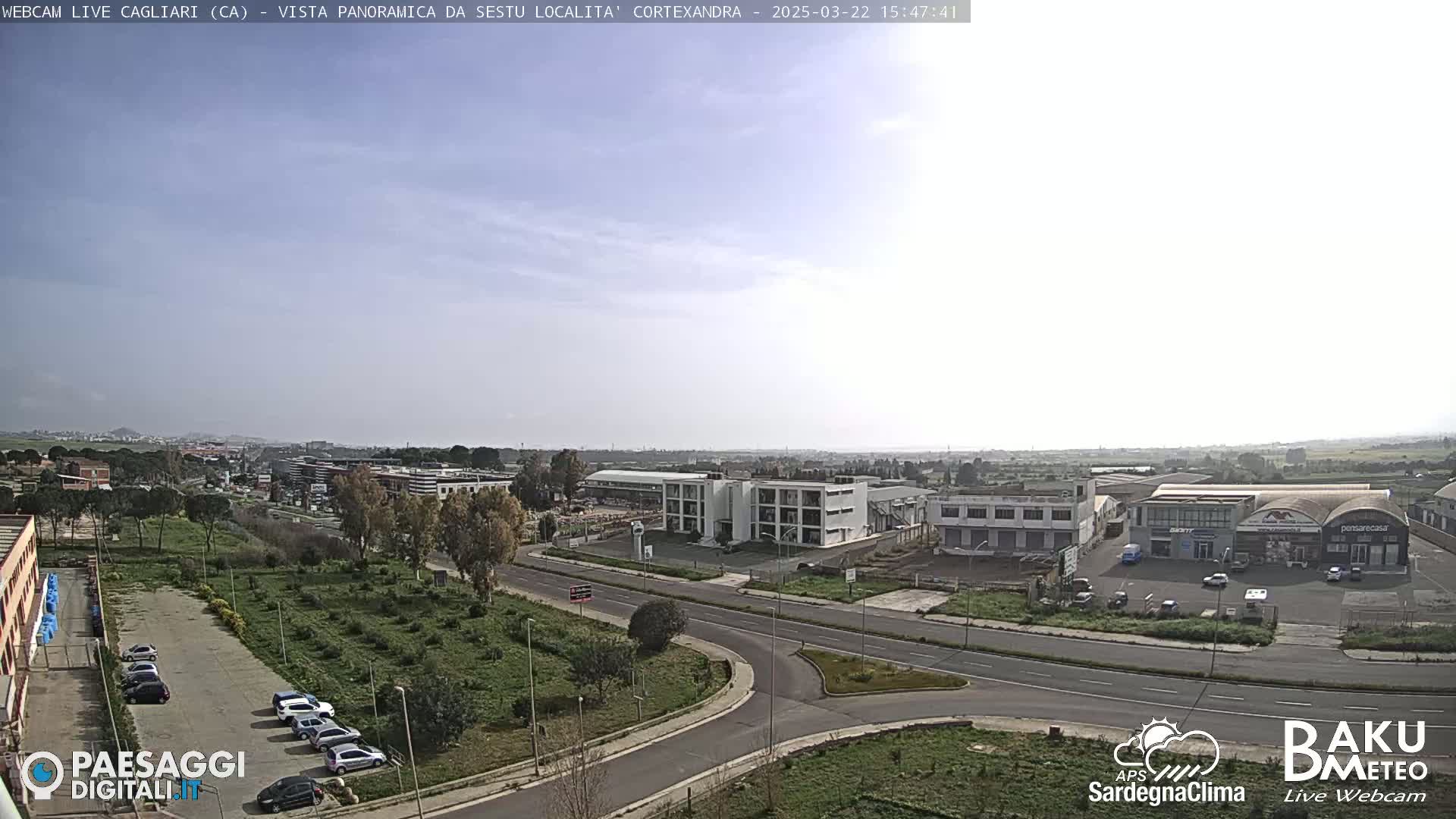 A mostly clear, sunny day shows a wide view of a city with industrial buildings, roads, and sparse vegetation.