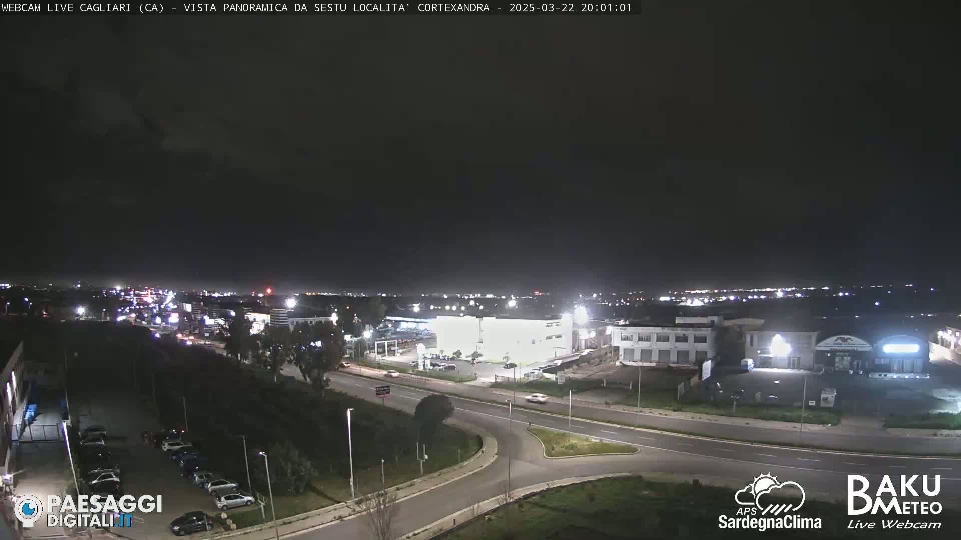 A nighttime, high-angle view of a road intersection and surrounding industrial area, with a city skyline visible in the distance under a clear, dark sky.