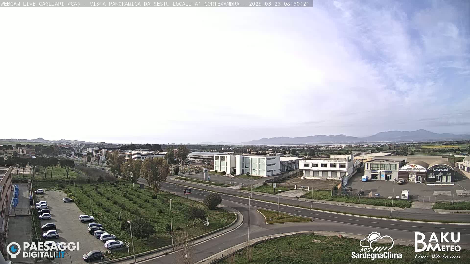 A mostly clear day reveals a panoramic view of a city with industrial buildings, roads, and a parking lot filled with cars, all set against a backdrop of distant mountains.