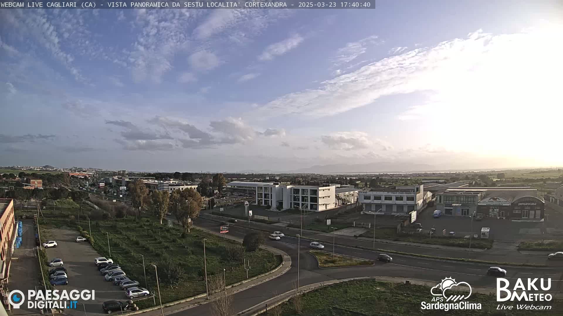 A partly cloudy afternoon view of a city with roads, buildings, and parked cars, overlooking a flat coastal area.