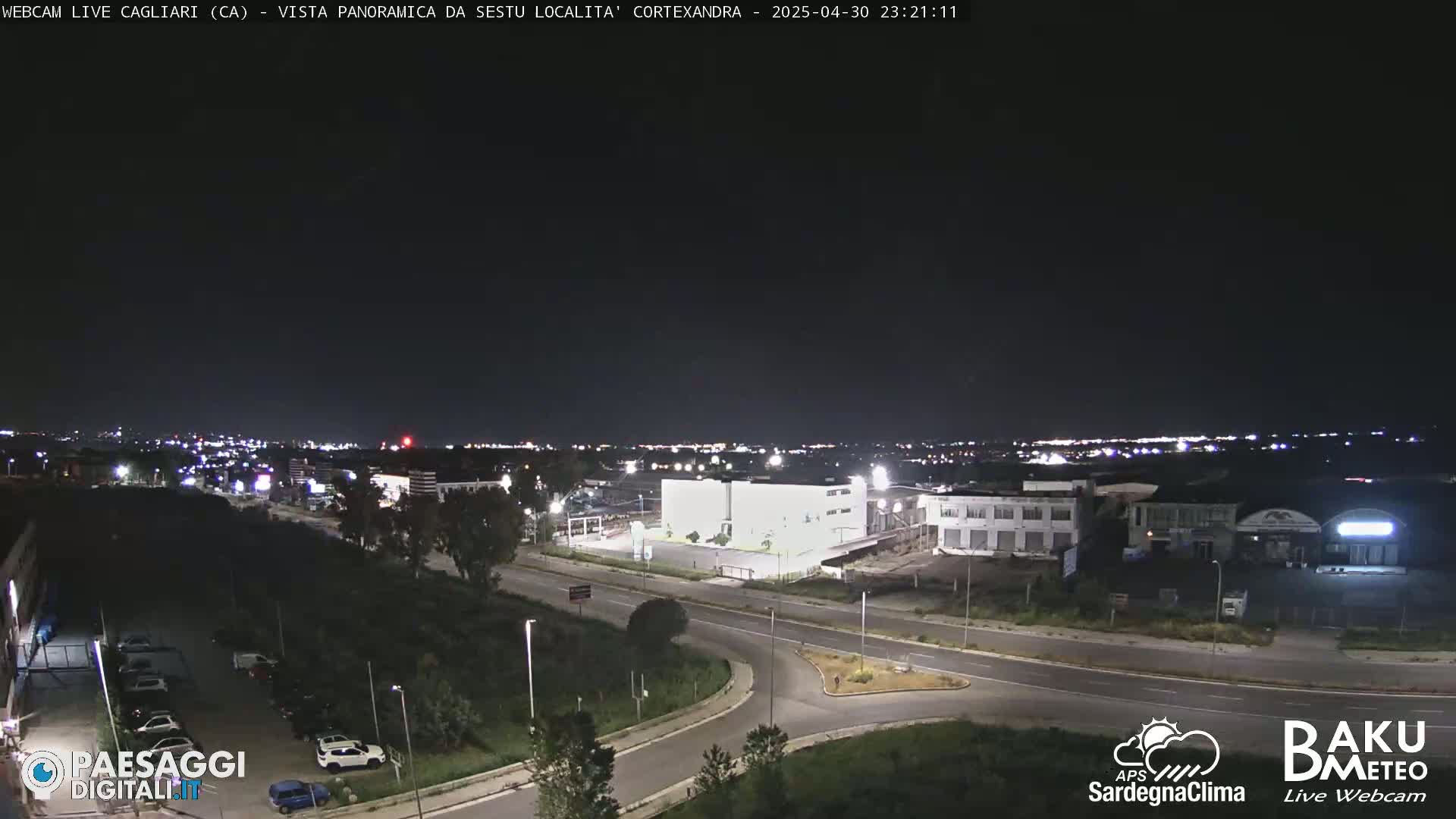 A nighttime, panoramic view of a city’s outskirts, showing roads, buildings, and sparse vegetation under a clear, dark sky.