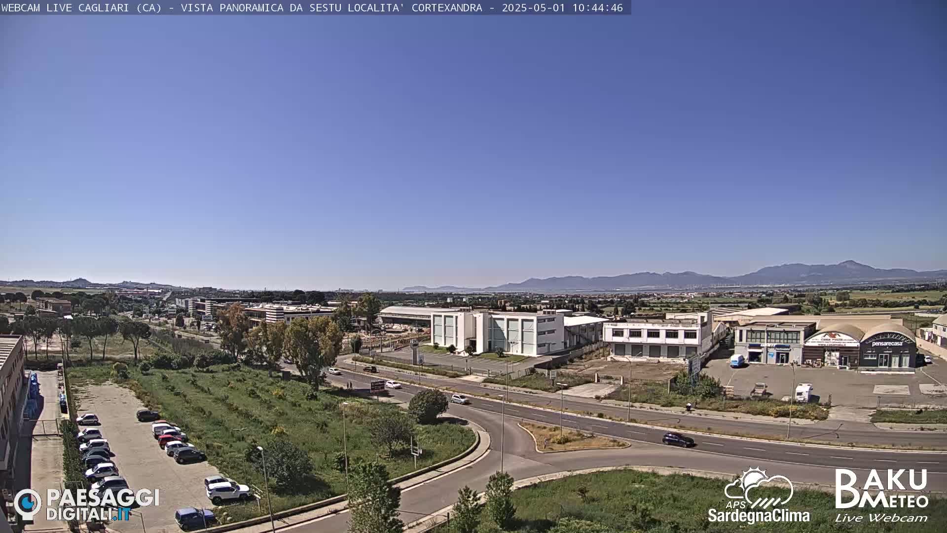 A clear, sunny day reveals a panoramic view of an industrial area with buildings and roads,  surrounded by green spaces and distant mountains.