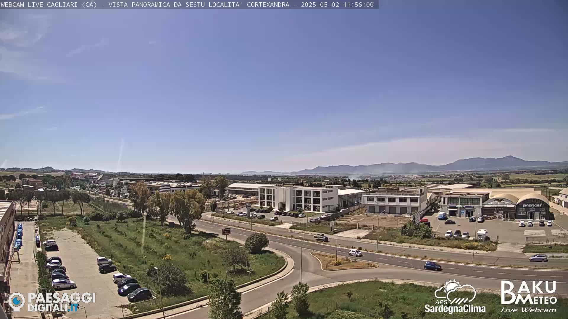 A sunny, high-angle view of a road intersection and industrial area, with distant mountains under a clear blue sky.