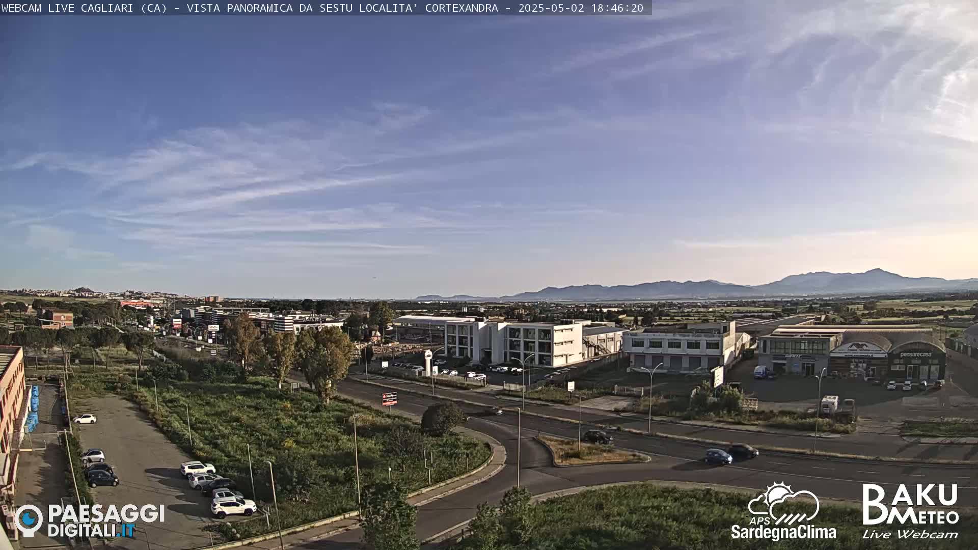 A clear, sunny day reveals an aerial view of a suburban industrial area with roads, buildings, and a few cars, all set against a backdrop of distant mountains.